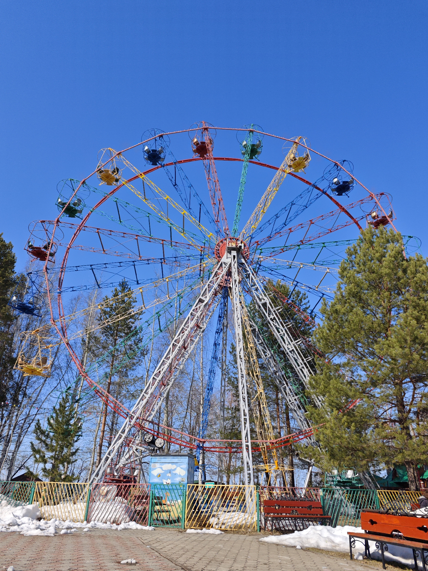 The image depicts a colorful Ferris wheel in a park-like setting under a clear, blue sky. The Ferris wheel has numerous cars, each painted a different color, attached to its perimeter. The supporting structure of the wheel is a mix of white, red, and other painted metal beams. The surrounding environment includes trees with varying degrees of foliage, patches of snow on the ground, a paved walkway, and park benches. A low fence with colorful, vertical bars borders the immediate area around the Ferris wheel. The scene is in the city of Chaikovsky, Russia. There are no visible people.