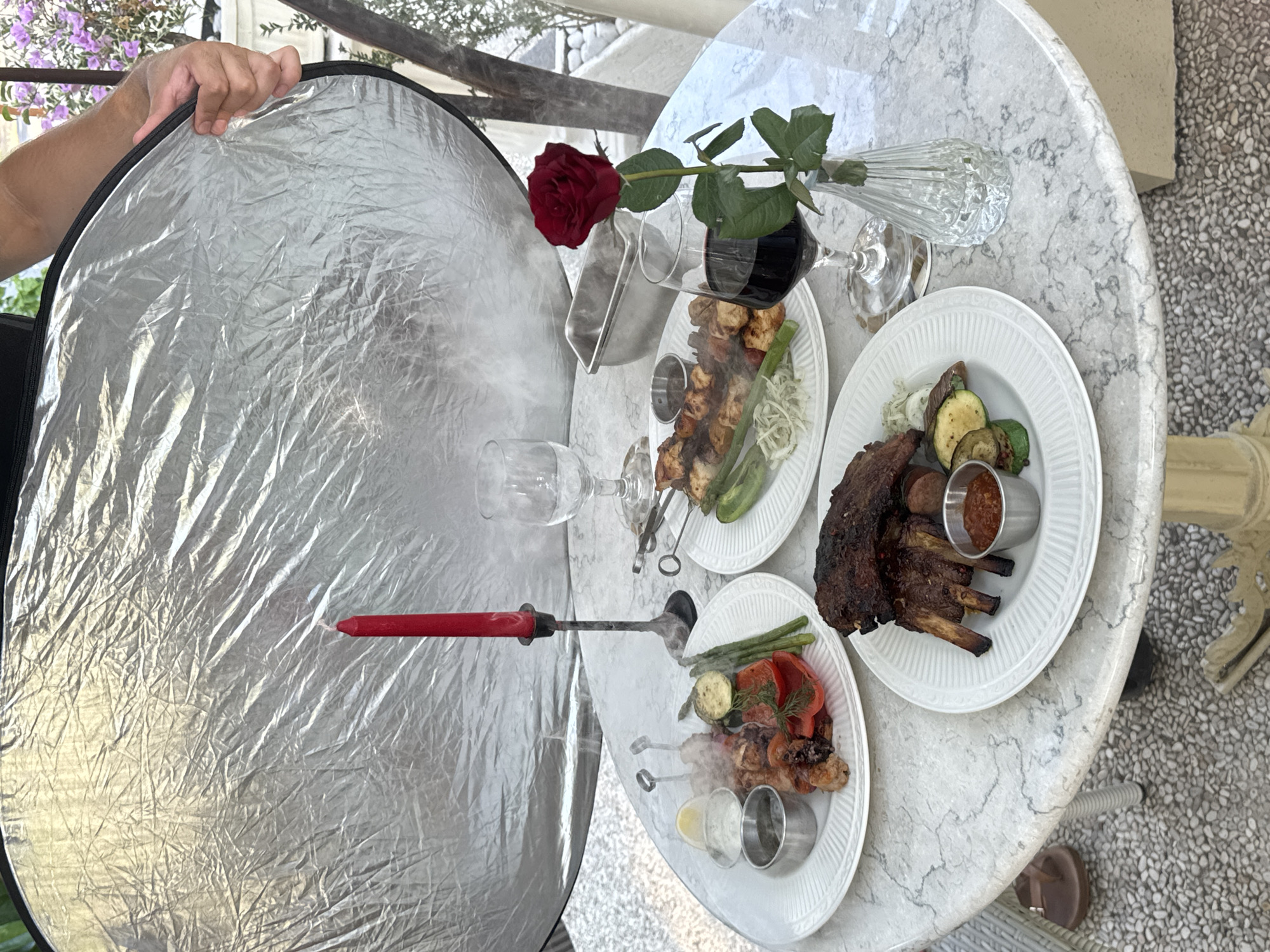 A photographer is using a large, silver reflector to bounce light onto a table set for a meal. The table, which appears to be made of white marble with gray veining, is set outdoors on a pebbled ground. On the table are two plates of grilled food, skewers of meat and vegetables, a glass of red wine, a single red rose in a small vase, and a red candle in a black candlestick. Steam is rising from one of the plates, suggesting the food is freshly cooked. The overall scene appears to be a staged food photography setup. The location in Karangasem, Indonesia is not depicted in the image.