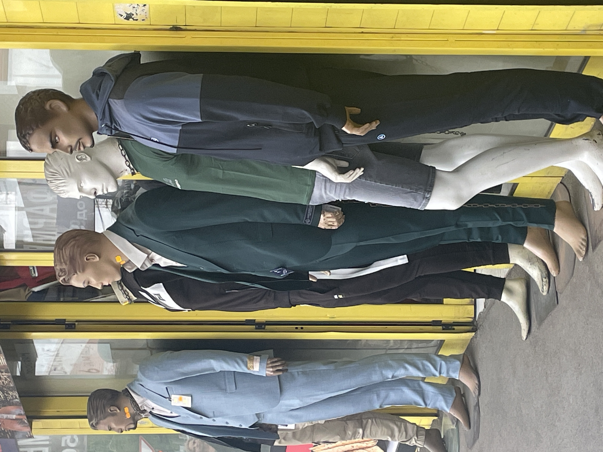 A shop window display in Belgrade, Serbia, features five horizontally arranged mannequins dressed in contemporary clothing. From top to bottom, the mannequins are wearing: a two-tone hooded jacket and dark pants; a green shirt and grey shorts; a dark green suit with a white shirt; a black jacket and dark pants; and a light blue suit. Each mannequin has a tag affixed to its chest. The backdrop includes reflections of the street and building exteriors.