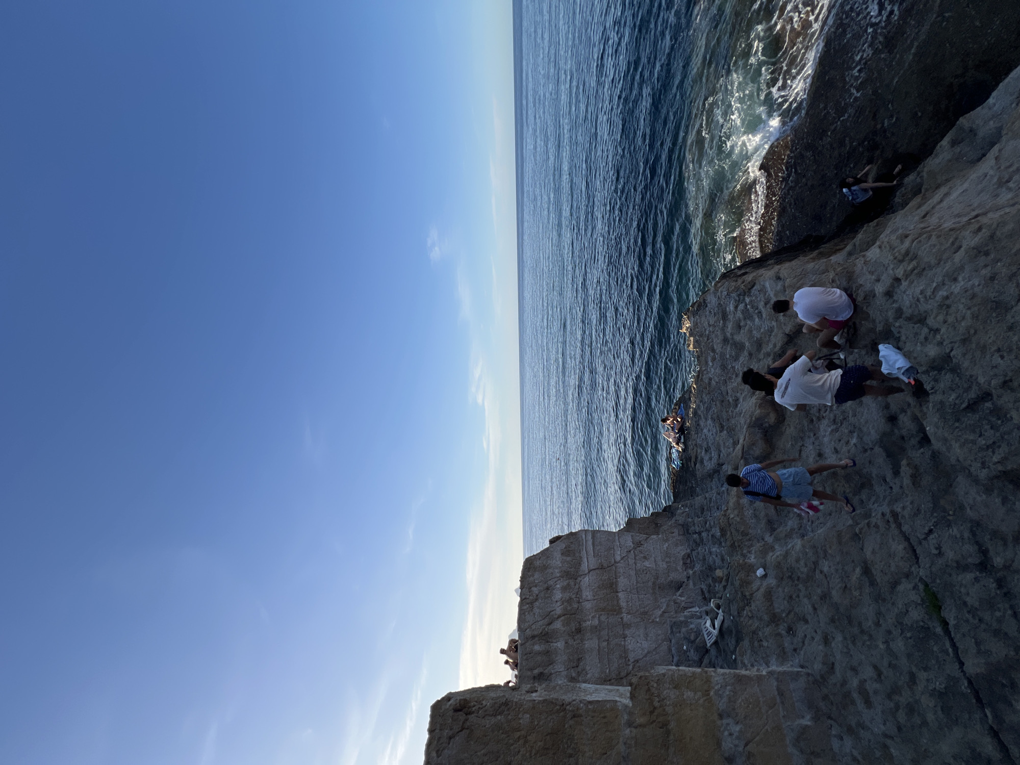 The image depicts a sunlit coastal scene in Sperone, Italy, showcasing a rugged rock formation bordering the sea under a clear blue sky with minimal high-altitude cloud cover.
The sky, occupying the upper portion of the frame, transitions from a lighter blue near the horizon on the left to a deeper azure towards the top right. The sun, positioned low on the left, casts a warm glow, creating some backlighting effects.
The Mediterranean Sea dominates the middle and right sections of the image, appearing as a deep blue body of water with discernible ripples and white-capped waves crashing against the rocky coastline in the lower right.
A large, multi-layered cliff, composed of light brown and gray sedimentary rock, rises on the left side of the image. Its sheer face and horizontal strata are clearly visible. Several individuals are situated on the flat top of this cliff, mostly silhouetted, appearing to be observing the view or resting.
Below the main cliff and extending towards the water, a series of darker, irregular rock formations and platforms create a natural terraced landscape. On one of these lower platforms, three individuals are present:
1. A person in a blue and white striped t-shirt, light blue shorts, and flip-flops, holding a red and white striped towel, stands facing away from the cliff and towards the sea.
2. To their right, a person in a white t-shirt and dark shorts is seen from the back, holding a white garment.
3. Next to them, a third person in a white t-shirt and pink or red shorts is seated on the rock, also viewed from the back.
Near these individuals, a white plastic cup and a white plastic bag or piece of litter are visible on the rocks.
Further down and to the right, a person with a backpack is seen navigating or climbing the steep, dark rocks near the water's edge. At the lowest accessible rocky platform, directly adjacent to the waves, two individuals are lying or sitting on a blue towel, appearing to be sunbathing or relaxing. The overall scene suggests a leisurely gathering in a natural coastal environment.