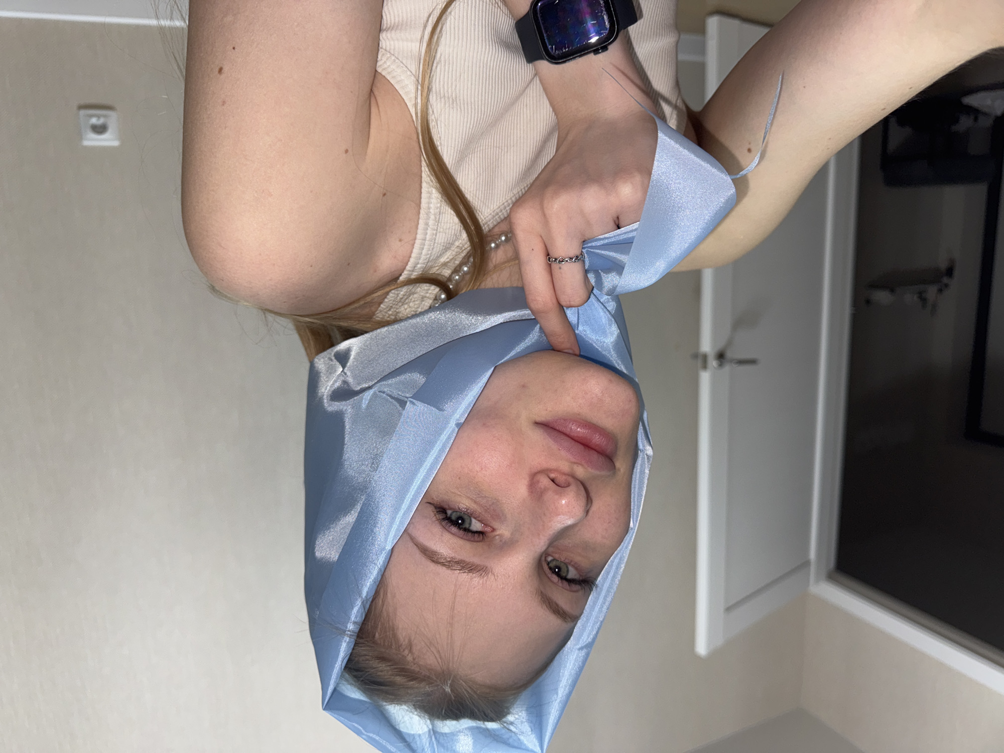 The image shows a young woman with blonde hair wearing a light blue satin scarf draped over her head and shoulders. She is looking towards the camera with a neutral expression. She is wearing a light-colored ribbed tank top and a ring on her left hand. A black smartwatch is visible on her right wrist. The background appears to be an indoor setting, with a beige wall, a white door frame, and a light fixture. The image is oriented vertically, with the woman's head at the bottom and her body extending upwards. There is no indication of the location being in the city of Perm, Russia.