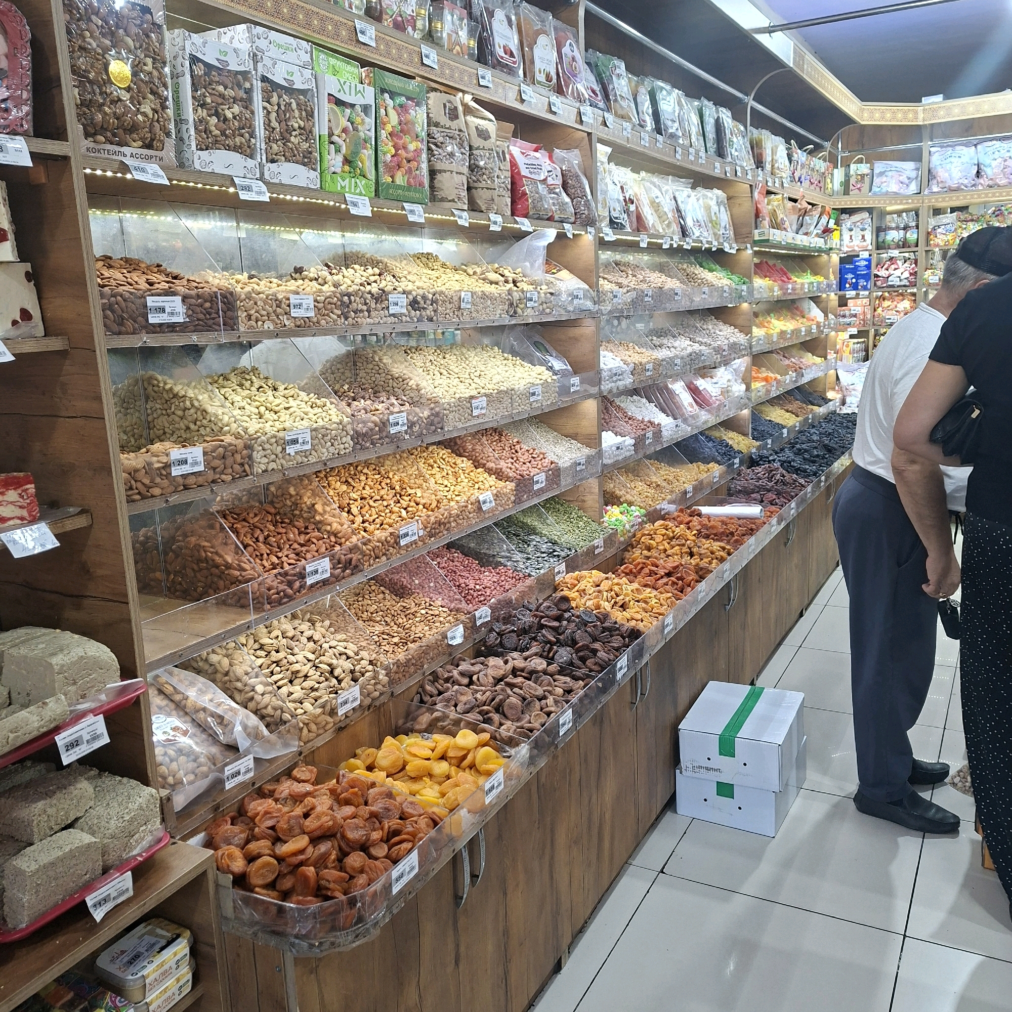 In an indoor market stall, a variety of nuts and dried fruits are displayed in clear plastic bins on wooden shelves. The products include almonds, cashews, pistachios, sunflower seeds, dried apricots, and prunes, among others. Some products are packaged in clear plastic bags. There are price labels on some of the bins. On the left side of the stall, there are blocks of halva. In the foreground, two people are partially visible, one wearing a white t-shirt and dark trousers, the other a black t-shirt and a black and white polka dot skirt. They are standing in front of the display. Two white boxes with green tape are stacked on the floor in the lower right corner. The lighting is artificial, and the overall environment suggests a retail setting.