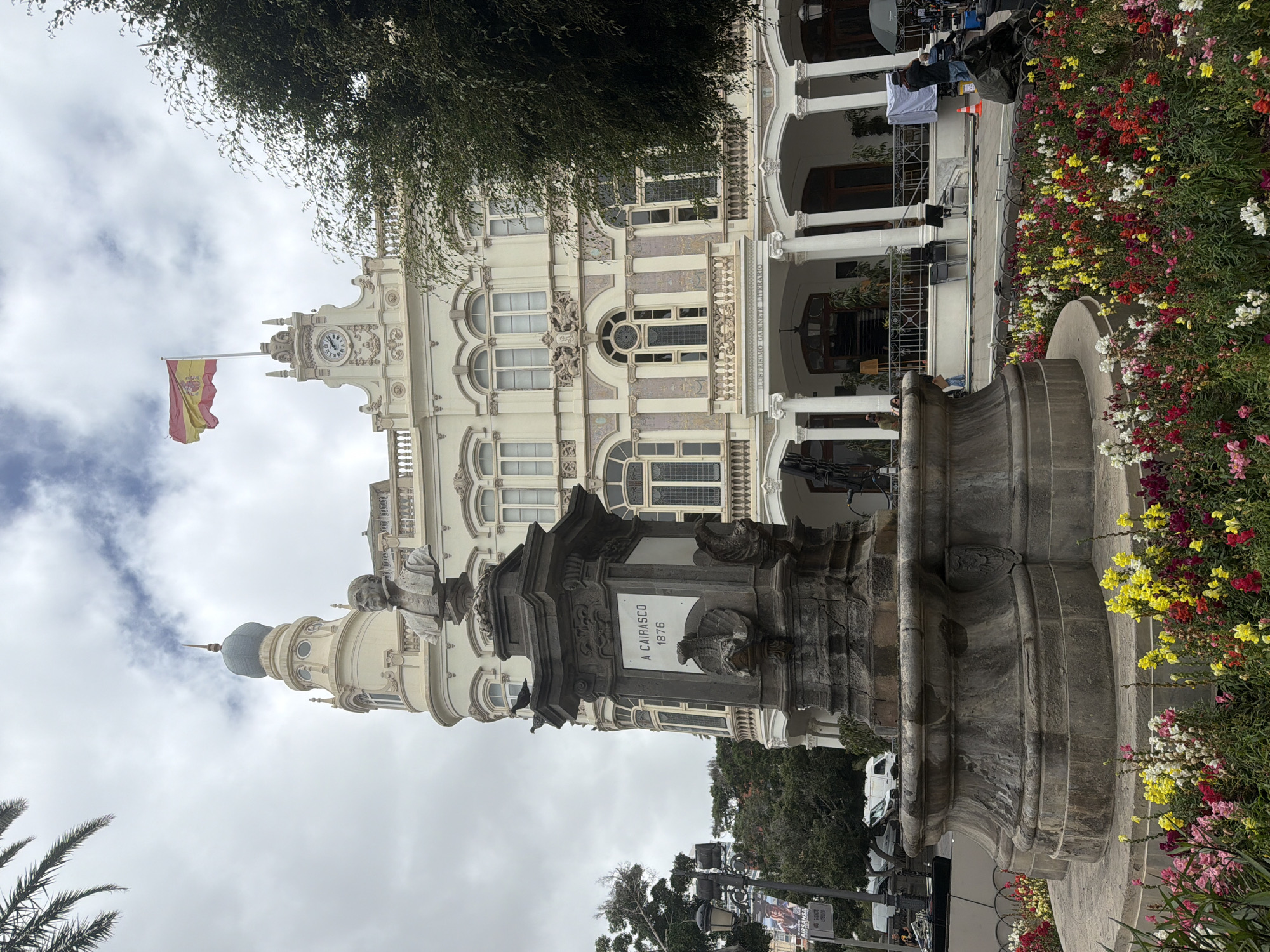 The image captures a view of the Gabinete Literario building in Las Palmas De Gran Canaria, Spain. In the foreground, a large stone monument with a bust of a man stands prominently. The monument is ornately carved and features a plaque that reads "A CAIRASCO 1876". Surrounding the monument is a vibrant flowerbed filled with a variety of colorful flowers, including red, yellow, pink, and white blooms.
The Gabinete Literario building itself is a grand, multi-story structure with a distinctive architectural style. It features a central dome with a spire and a Spanish flag flying from its rooftop. The facade of the building is adorned with decorative elements, arched windows, and balconies. A portico with classical columns leads to the entrance.
In the background, trees and palm fronds are visible. Several people are present in the scene, some standing near the building's entrance, while others are on the street in front of it. Some appear to be involved in activities related to filming or photography, with equipment such as cameras and lighting rigs visible. The sky is overcast, suggesting a cloudy day. The overall scene portrays a public square with a notable historical building and a monument, bustling with activity.