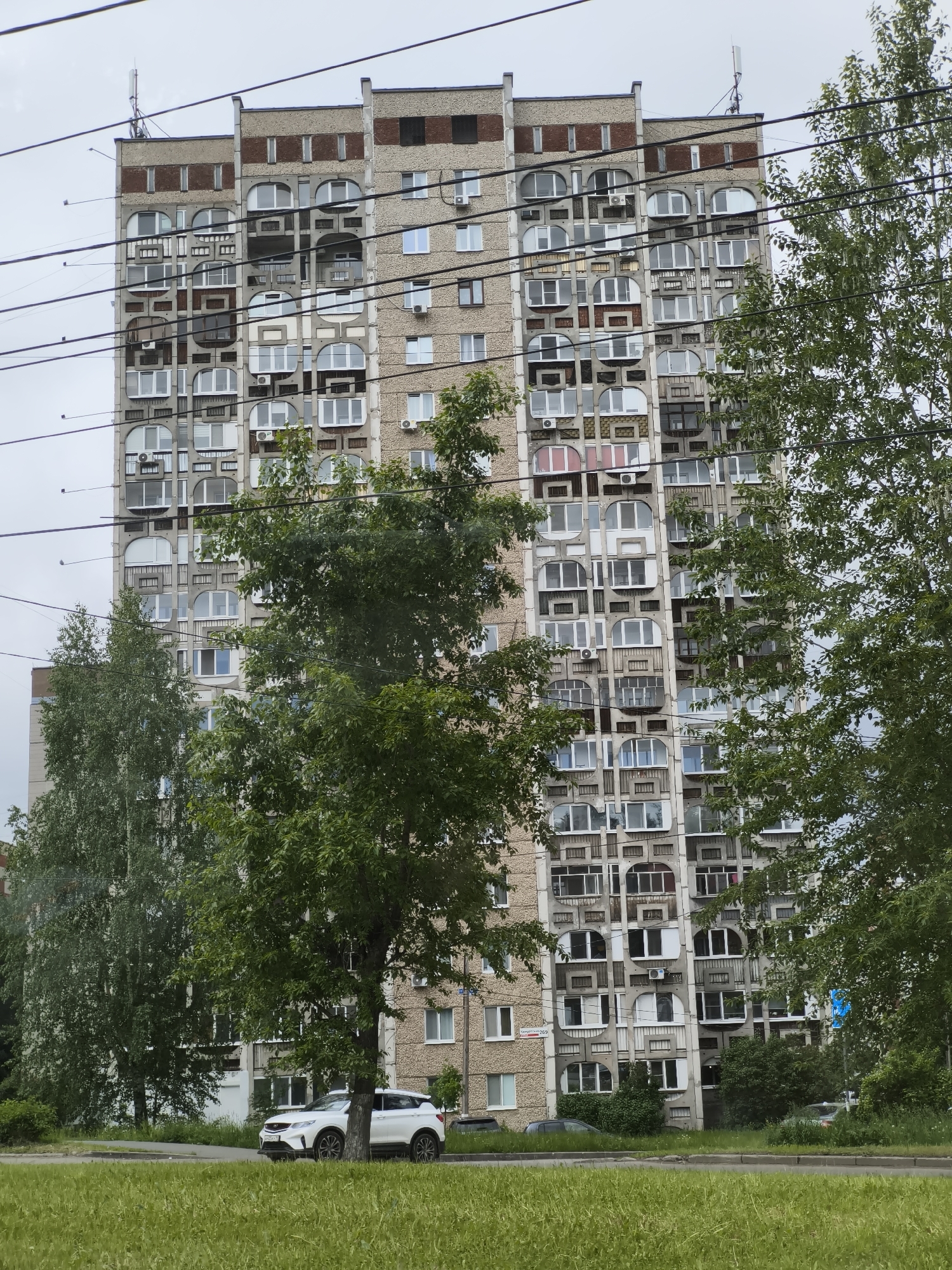 The image captures a tall, multi-story residential building in Izhevsk, Russia, under an overcast sky during daylight hours. The building exhibits a distinctive Soviet-era architectural style, possibly brutalist, characterized by a complex facade featuring a multitude of windows in various shapes, including many prominent arched designs interspersed with rectangular ones. The exterior appears to be concrete, with some sections showing exposed brick, and numerous air conditioning units are affixed to the walls.
In the foreground and midground, several green deciduous trees partially obscure the view of the building, adding natural elements to the urban landscape. A strip of green grass runs along a paved road in the lower part of the frame. A white SUV is parked on the side of the road, and other vehicles are visible further down near the building. Multiple black power lines stretch horizontally across the image, running in front of both the trees and the building. Antennas or communication equipment are visible on the building's roof.
On the lower left side of the building, a sign with the text "Универмаг 200" is visible, which translates to "Department Store 200". The white SUV's license plate displays "У223ОХ 18", where "18" is the regional code for the Udmurt Republic, confirming the location as Izhevsk, Russia. The scene depicts a static urban environment with no specific activity or event taking place.