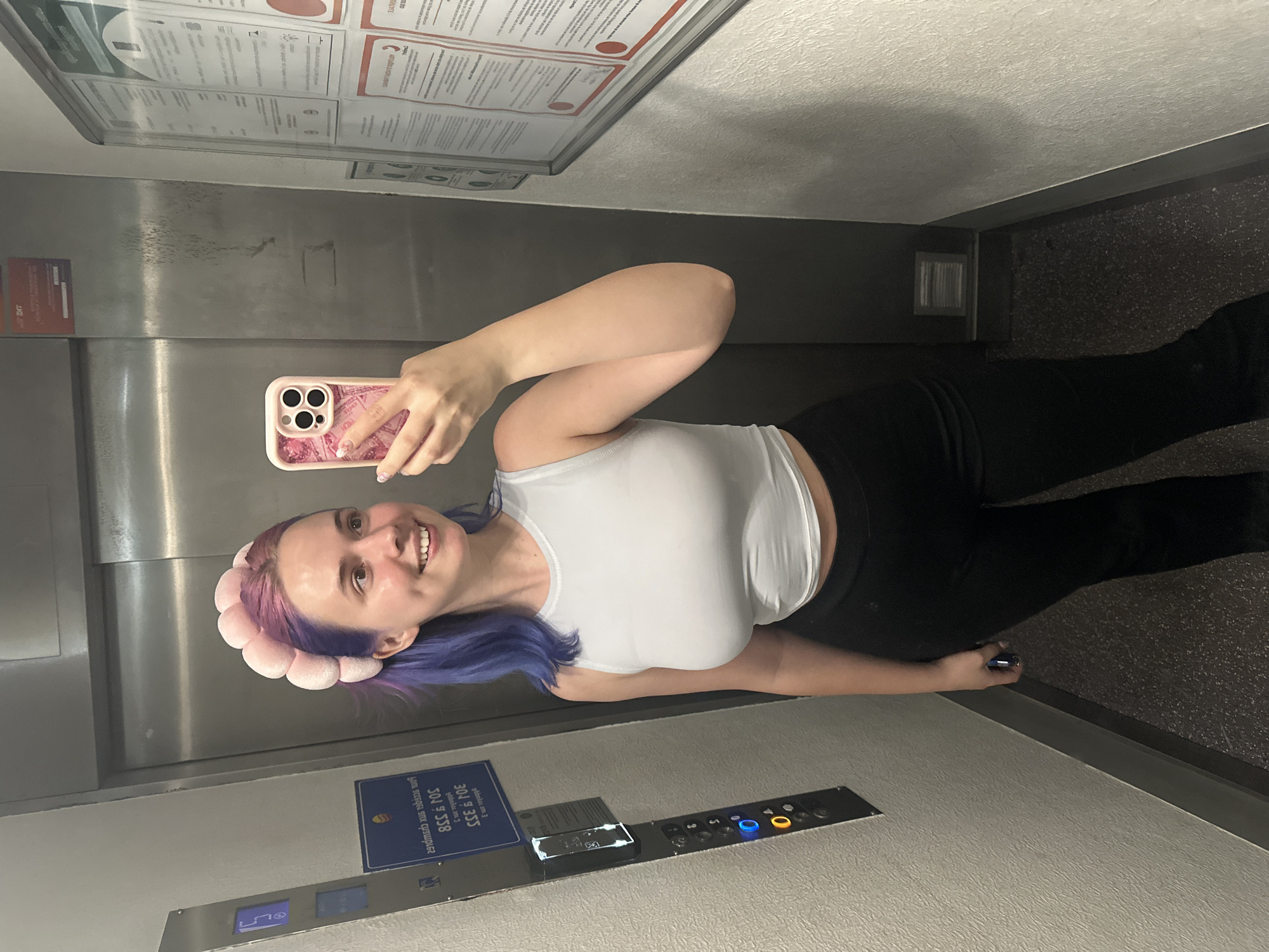 A woman stands in an elevator, holding a pink mobile phone, presumably taking a selfie. She has light skin, purple hair, and wears a white tank top and black pants. She also sports a pink flower-like headband. The elevator features metal doors, a control panel with numbered buttons and a digital display, and a sign with text in French. The elevator’s interior appears to have carpeted flooring. Given that the sign on the wall has the telephone number “304 9 335," the location is confirmed to be in Ramonville-Saint-Agne, France.
