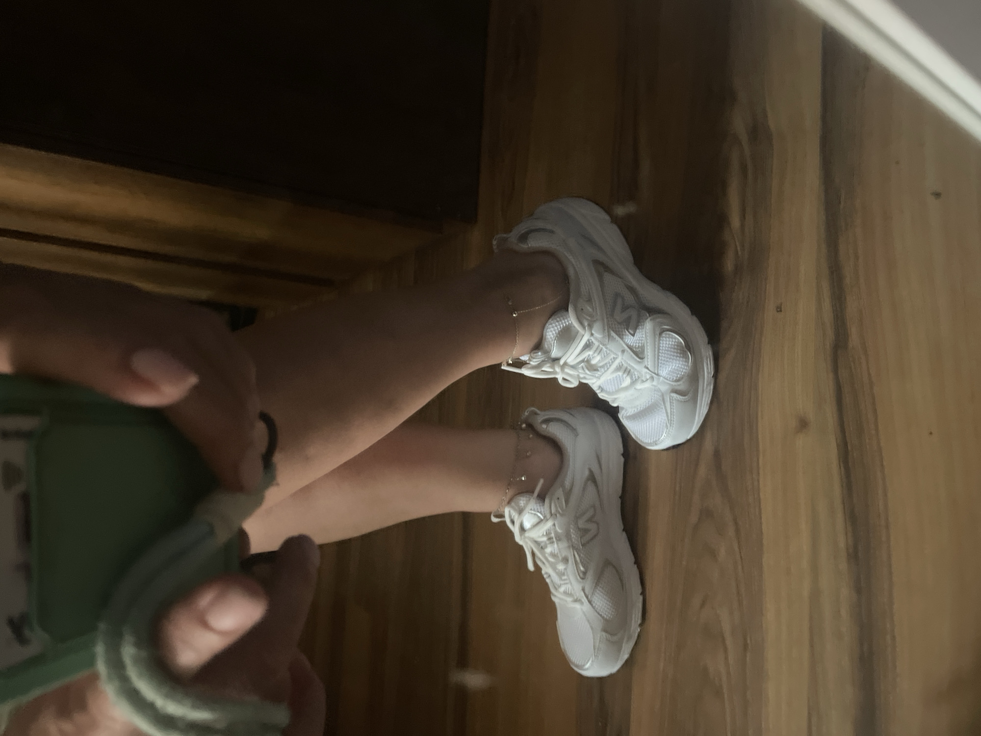 The image shows a close-up of a person's legs and feet, clad in white athletic shoes. The shoes have silver accents and appear to be laced. Each ankle is adorned with a delicate silver anklet. The person is standing on a wooden floor, which has a warm, polished finish. In the background, a dark wooden door is visible, adding a sense of depth to the scene. A soft, green item, possibly a cloth or part of a phone case, is held in the person's left hand, partially obscuring their legs. The lighting is dim, creating shadows and highlighting the texture of the wooden floor. The image is cropped to focus on the lower extremities and the shoes, suggesting an emphasis on footwear or personal style. The location in Kadıköy, Turkey is not visually represented in the image provided.