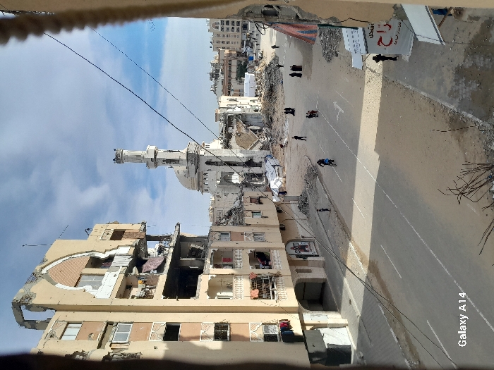 The image displays a street scene in Khan Yunis, Palestinian Territory, captured from an elevated vantage point, looking down a paved street under a partly cloudy sky.
On the left, a multi-story building exhibits extensive structural damage, including partially collapsed upper floors, exposed interiors, and missing facade sections, though laundry is visible hanging from balconies on its lower, less damaged levels. In the central background, a prominent mosque appears severely damaged, with its minaret and dome compromised, surrounded by significant debris piles.
The street itself is a two-lane asphalt road with faded white markings, flanked by dirt and rubble. Scattered across the street are approximately a dozen individuals. One person is observed riding a bicycle down the center lane. Several pedestrians, some in small groups and others individually, walk along the street or stand near the roadside. On the right side, a partially visible storefront with Arabic script is present. In the distant background, additional damaged structures and areas with apparent makeshift shelters or debris are visible. Overhead wires traverse the scene.