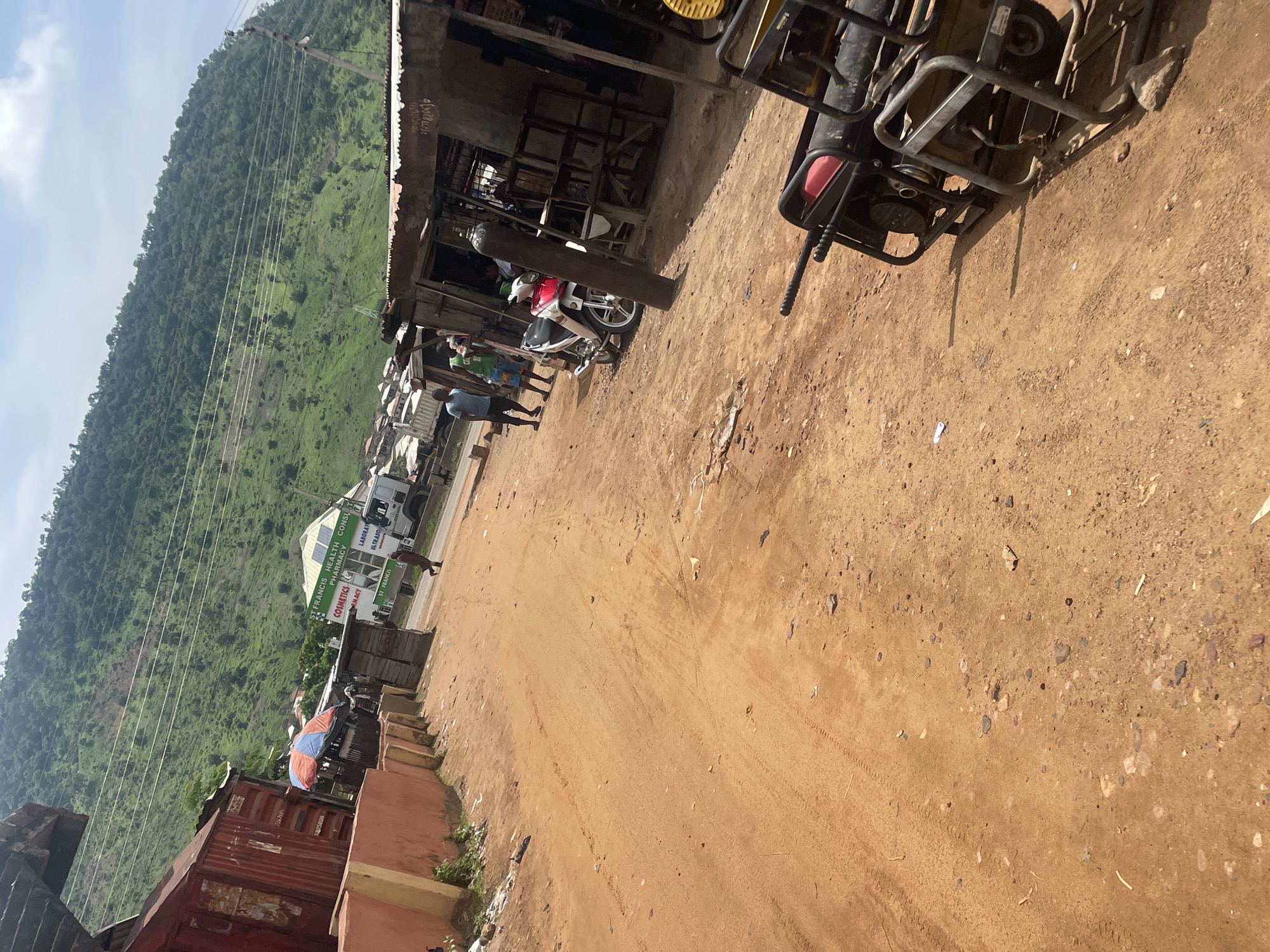 This image depicts a street scene in Lokoja, Nigeria, characterized by a dirt road and a backdrop of a lush green, forested hillside. On the left side of the road, a large, red shipping container sits against a rough, orange-colored wall. Further down the road, there is a building with signs that read "ST. FRANCIS HEALTH CONSULT PHARMACY" and "LABORATORY ULTRASOUND". Several individuals are present, with two men standing near a motorcycle and a third person walking on the road in the distance. Several makeshift stalls or shops line the right side of the street, constructed from corrugated metal and wood. Parked on the side of the road are two generators, one black and red and another yellow and silver. The overall impression is of a vibrant, bustling, yet somewhat rustic, urban environment within a natural setting.
