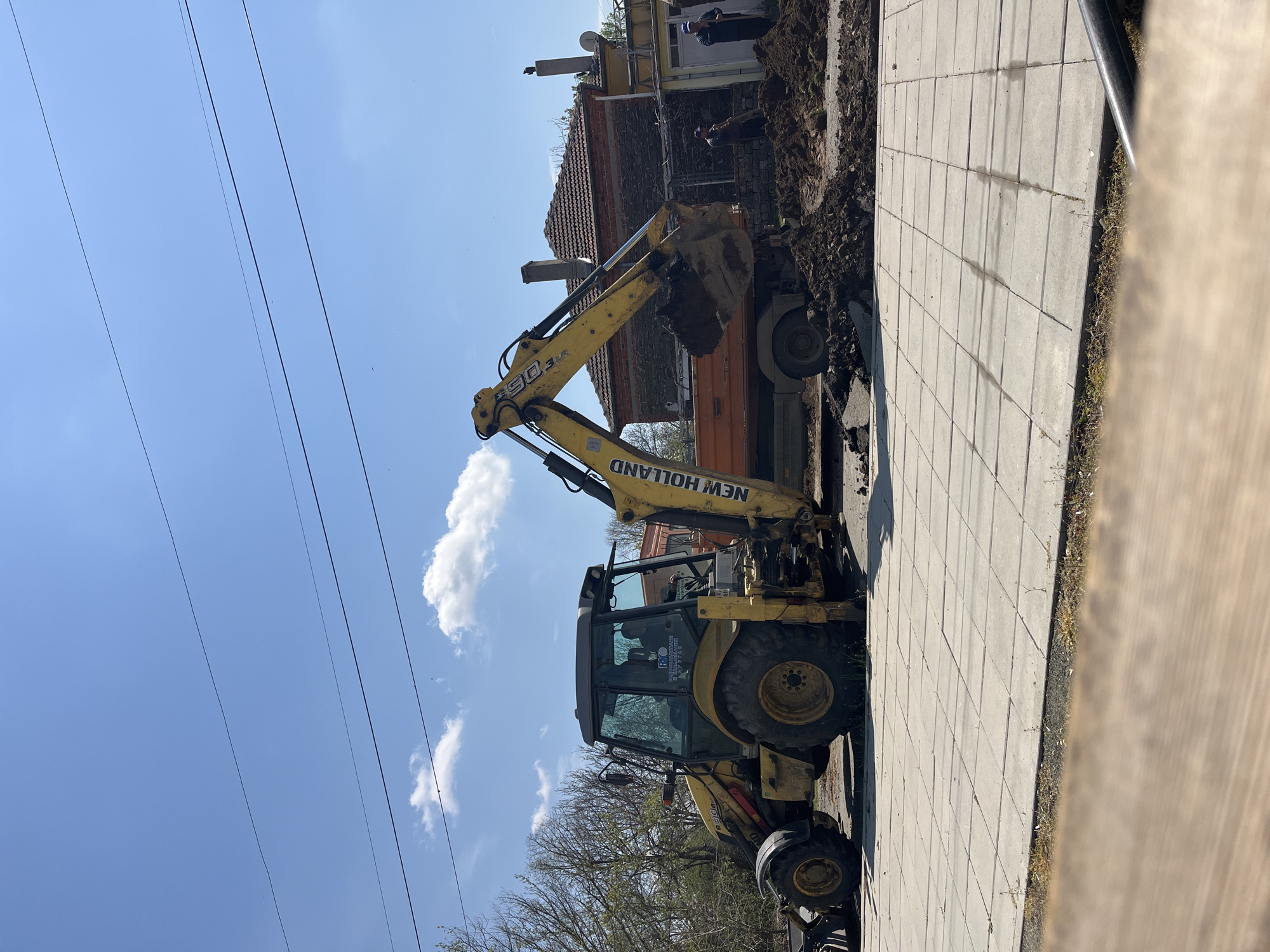 The image captures an outdoor construction or excavation scene in broad daylight, likely in Lozarevo, Bulgaria. A large yellow New Holland backhoe loader dominates the left and center of the frame, actively engaged in digging or loading. Its front bucket is full of dark earth and positioned above the bed of a partially visible orange dump truck, indicating the transfer of excavated material.
Two individuals are discernible near a two-story building in the background. One person stands upright near an opening in the building, while another appears to be bending over in the adjacent excavated area. The building itself has a red tiled roof, brick elements, chimneys, and multiple satellite dishes, suggesting it is a residential or commercial structure.
To the right of the excavation site, a neatly laid grey paved sidewalk, made up of square slabs, stretches horizontally. Beyond the sidewalk, a narrow strip of grass and a light-colored wooden fence or structure are visible on the far right. The ground around the machinery is disturbed, with piles of dirt from the excavation.
The sky above is a clear, bright blue with scattered white, fluffy clouds, suggesting sunny weather. Several dark utility lines traverse the upper portion of the sky. In the lower left, the bare branches and emerging leaves of trees can be seen.
Visible text on the excavator includes "NEW HOLLAND" and "B90 BLR" on its arm, as well as a sticker on the cab displaying "БПС" (BPS) and other unreadable Cyrillic script. The scene portrays ongoing outdoor work in what appears to be a typical town or village environment.