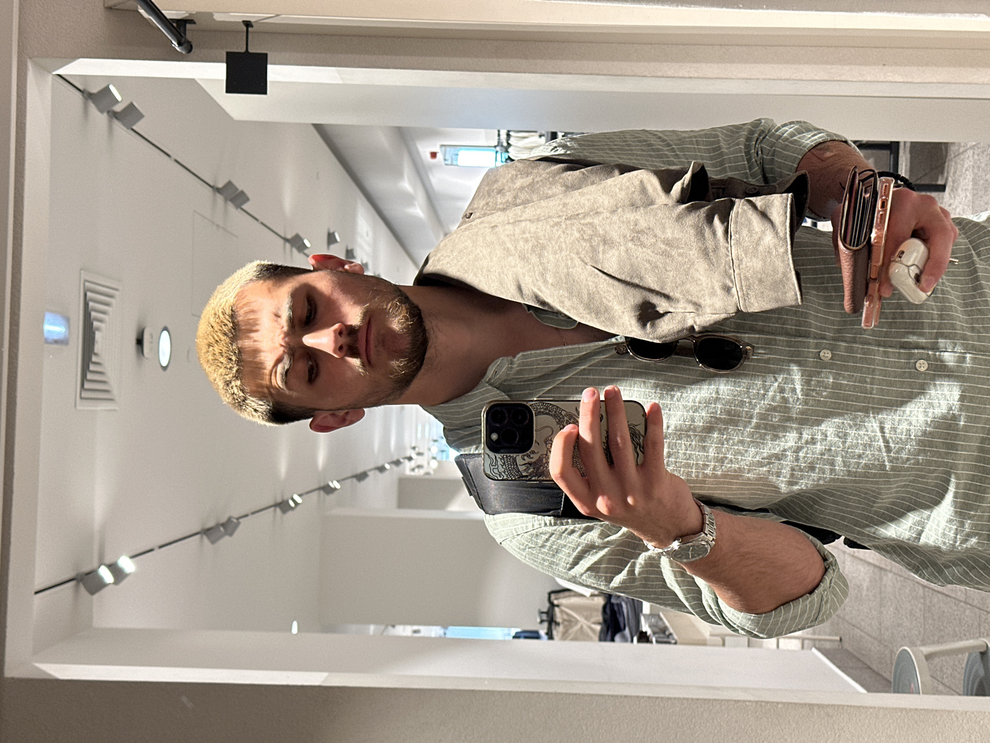 A man is standing indoors, likely in a clothing store, taking a selfie in a mirror. The man has blond hair, a beard, and is wearing a striped button-down shirt with a tan jacket draped over his shoulder. He is holding a phone in his right hand and accessories such as sunglasses, a wallet, and earphones in his left hand. In the background, there are racks of clothing and overhead lighting that suggest a retail environment. Based on the interior design, this could be a branch of a popular international chain clothing retailer, such as Zara or H&M. Sofia, Bulgaria isn't immediately discernible from the image without further context.