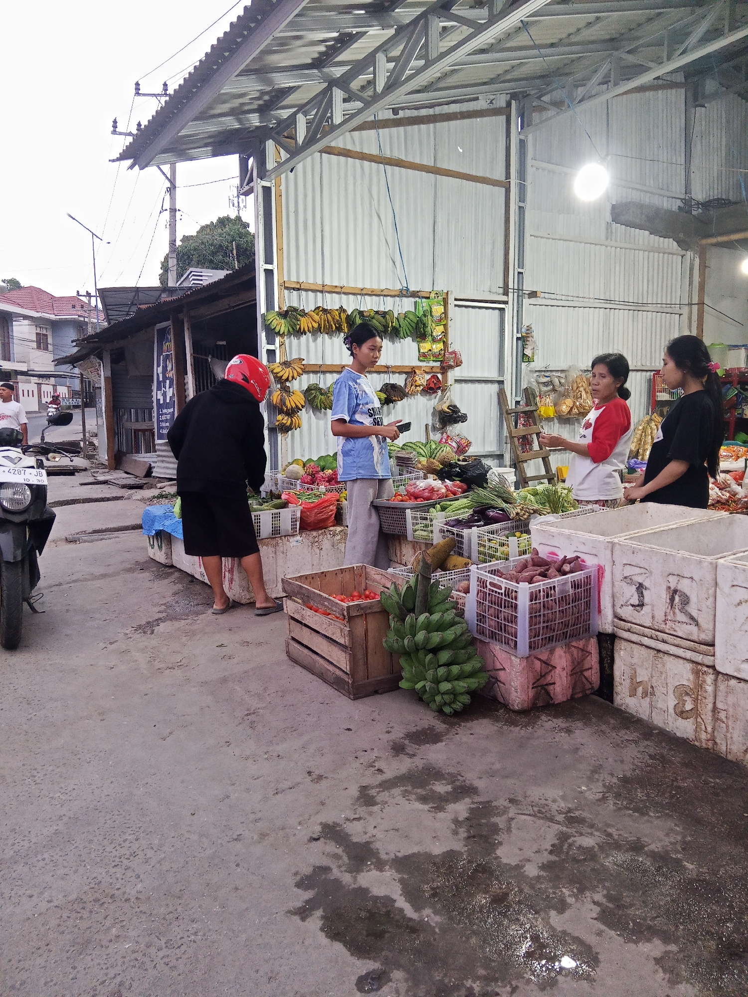 The image depicts a street-level market scene in Makale, Indonesia, likely located near a road. The foreground shows a concrete ground with some wet patches and a partial view of a motorcycle on the left. A person wearing a black hoodie and shorts, with a red helmet, is standing facing away from the camera, appearing to be interacting with the market stall. To the right, a market stall is set up under a corrugated metal roof supported by a metal frame. The stall displays a variety of fresh produce, including bananas hanging from the ceiling, crates of vegetables like eggplants and cucumbers, watermelons, and other fruits and vegetables in plastic crates and wooden boxes. Three individuals are visible at the market stall. One person, wearing a blue t-shirt, is standing on the left, holding a mobile phone. In the center, a woman in a red and white t-shirt is talking to another woman on the right, who is wearing a black t-shirt. The background shows buildings and trees, suggesting an urban or semi-urban environment.