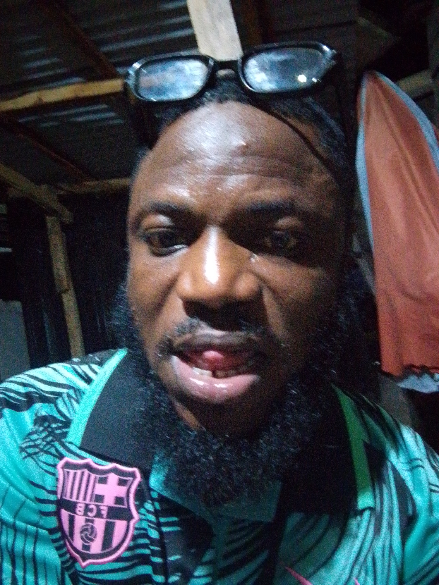 A close-up selfie photograph shows a man with dark skin, a black beard, and black-framed glasses perched on his forehead. He is wearing a green and black striped soccer jersey with a pink Barcelona team crest on the left side of his chest. His mouth is slightly open, revealing a glimpse of his tongue and teeth. The background is dimly lit, suggesting an indoor location. Wooden beams of the roof are visible above the man. A draped piece of fabric hangs to the right. No specific information within the image indicates a location in Ajaokuta, Nigeria.
