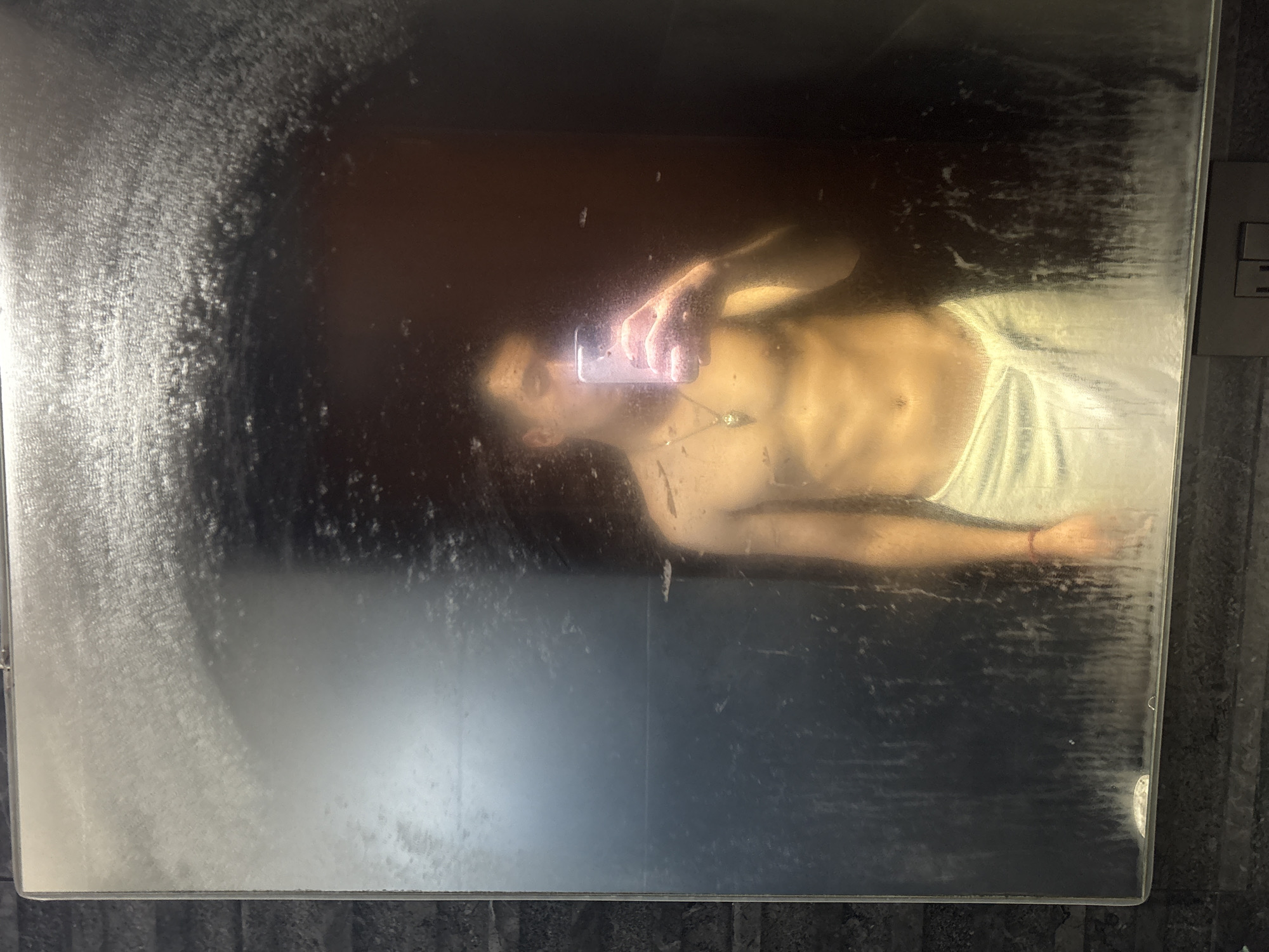 The image captures a selfie being taken in a highly condensed or wet mirror, likely in a bathroom setting in San Bernardino Tlaxcalancingo, Mexico.
The primary subject is a person, appearing to be male, reflected in the mirror. They are shirtless, with a light-colored towel or wrap around their waist. Their skin has a warm, golden hue, possibly from artificial lighting. The person is holding a white smartphone with their right hand, angling it towards their reflection to take the photo. A dark pendant or necklace is visible around their neck. Due to the mirror's condition, the person's facial features are very indistinct and hazy.
The mirror or glass surface is heavily covered in water droplets, condensation, and streaks, which significantly obscure and distort the reflection. This suggests a steamy environment, possibly just after a shower. The left side of the mirror shows dense, uneven streaking, while the bottom edge also has visible watermarks.
In the background, to the right of the reflection, a light-colored electrical outlet with two horizontal slots is mounted on a dark, textured wall. The edges of the mirror are visible, framed by this dark wall. The overall scene conveys a private, domestic moment. No discernible text is visible anywhere in the image.