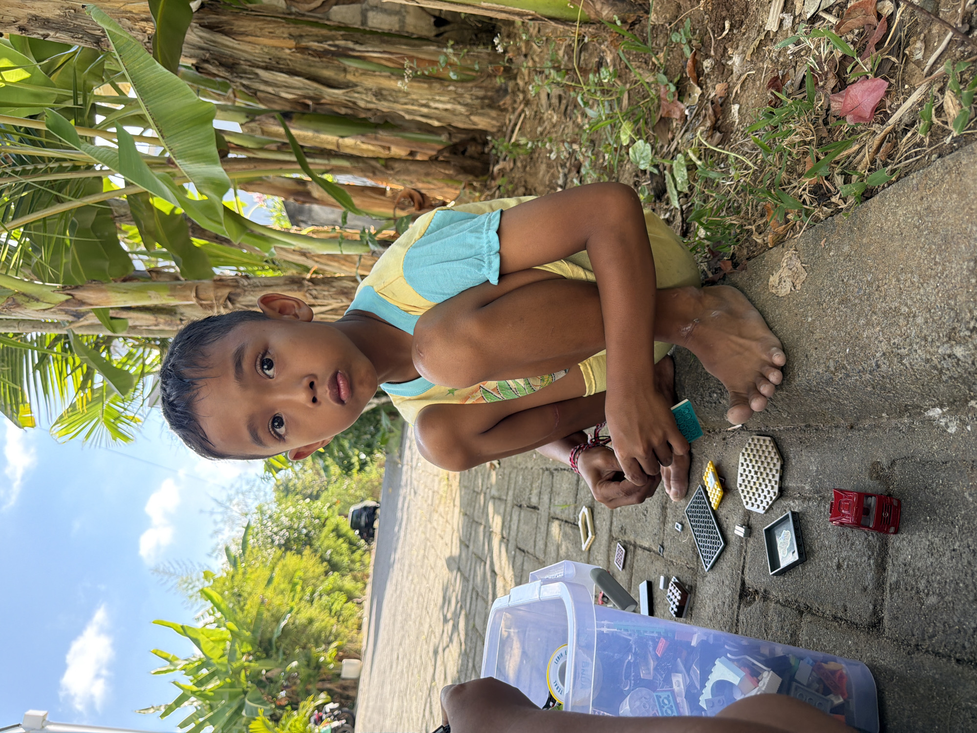 This is a medium-shot, eye-level photograph taken outdoors, likely during the day. The main subject is a young boy, seen from the chest up, squatting on a paved surface. He is looking directly at the camera with a neutral expression. He is wearing a yellow t-shirt with light blue sleeves.
Around the boy are various toys, including building blocks, a small red toy car, and other small construction pieces scattered on the ground. A clear plastic storage bin filled with more building blocks is also visible in the foreground. The boy's hands are interacting with some of these toys. His bare feet are visible on the ground.
The background includes a rough, textured wall or tree trunks with dry leaves and sparse greenery, suggesting a natural or semi-urban outdoor environment. In the upper left corner, the bright green leaves of what appear to be banana trees are prominent, and the sky with some clouds is visible. The paved surface is made of interlocking gray bricks.
The visible text in the image, on a yellow sticker inside the plastic bin, reads "LEBIH TEBAL BNDA". The overall scene suggests a child engaged in playing with building blocks outdoors, possibly in a backyard or a common area. The lighting and shadows indicate it is daytime, with the sun casting shadows on the ground.