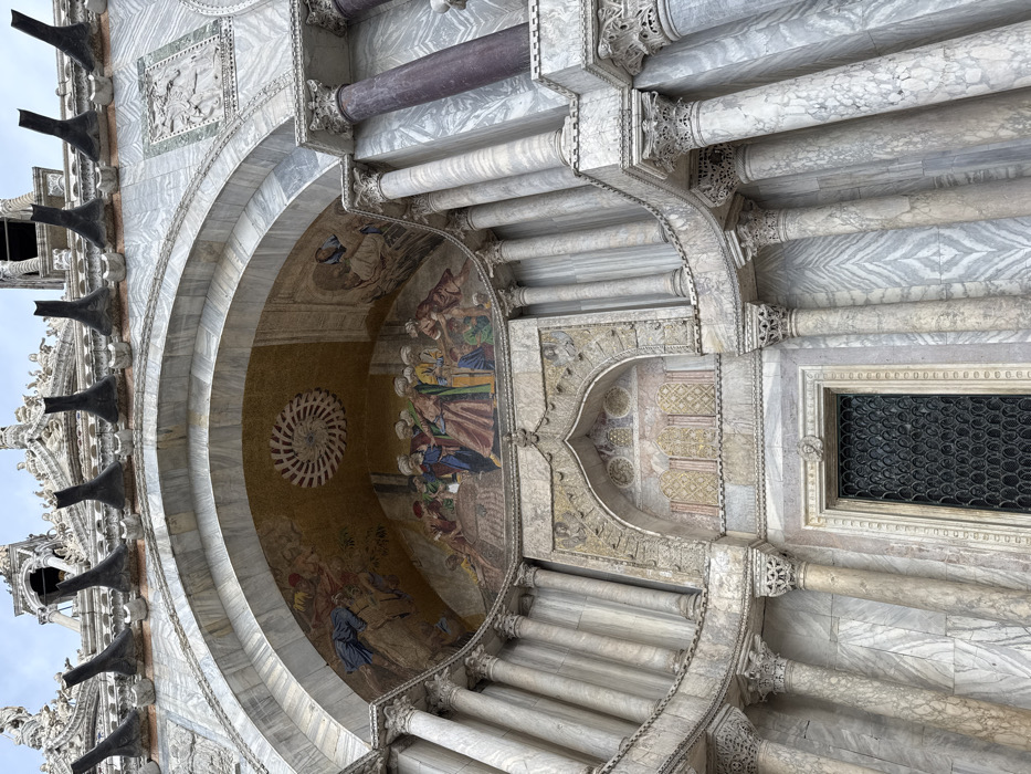 The image displays an upward-angled, detailed view of a section of St. Mark's Basilica in St. Mark's Square, Venice, Italy. The central focus is a large, multi-layered archway carved into the building's facade, constructed predominantly of white and grey veined marble.
Within the main arch, a vibrant mosaic panel is visible. The mosaic's upper central portion features a circular, golden spiral pattern. Below this, the mosaic depicts multiple figures. On the left, a figure mounted on what appears to be a horse is surrounded by other individuals. The lower and central mosaic sections illustrate a gathering of approximately ten robed figures, dressed in various colors including red, blue, green, and yellow, clustered around a sarcophagus or altar-like structure, engaged in the act of transporting a body.
The archway is deeply recessed and lined with multiple slender columns, featuring ornate Corinthian capitals. These columns are primarily white or light grey marble, with one prominent, darker, possibly porphyry, column visible on the upper right side. Above the arch, along the building's cornice, a series of seven dark, horn-shaped or bell-shaped decorative elements project upwards.
Below the large mosaic and archway, a smaller, intricately carved arch frames a dark double-leaf doorway. The doorway is covered by a patterned metal grille composed of numerous small, interlocking circular or scale-like elements. The surrounding marble facade exhibits strong natural veining patterns, with some sections featuring distinct chevron or herringbone designs. The entire structure is bathed in natural light under an overcast sky.