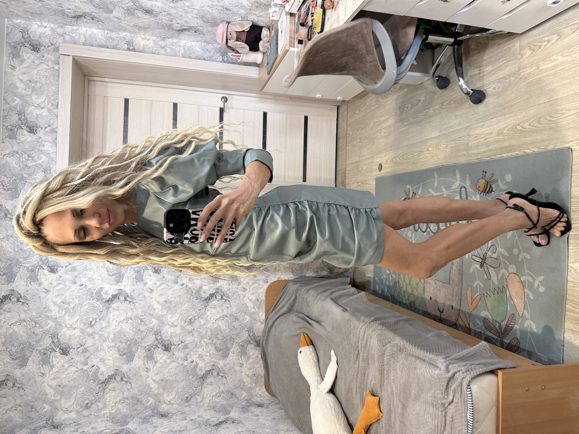 A mirror selfie of a female in an unknown location in the city of Unknown, Unknown. The woman has long blonde braided hair, wearing a light green ruffled dress and black strappy heels, holding a cell phone with decorated case in her right hand. The background shows a room with light grey patterned wallpaper, a partially visible white door with dark vertical stripes, a child's bed with a grey blanket and a white and orange duck plush toy, a children's rug with an illustration of animals, a desk with drawers, a brown chair and some items on the desk.