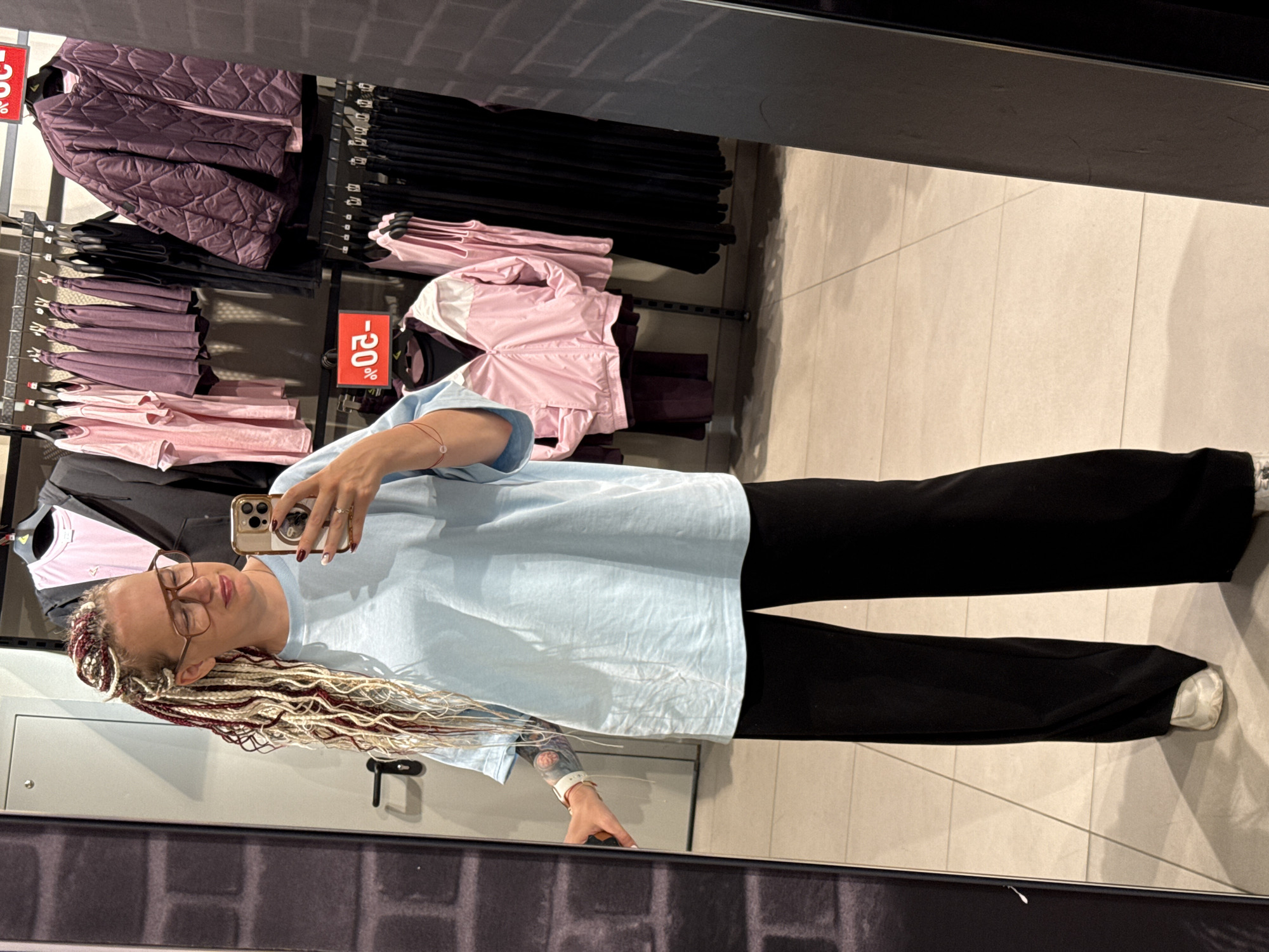 A woman with blonde and burgundy braided hair, wearing glasses, a light blue oversized t-shirt, and black wide-leg pants, is taking a mirror selfie in what appears to be a clothing store. She is holding a smartphone with a gold case and has a white smartwatch on her left wrist. The background shows racks of clothing, including pink and purple tops, a purple quilted jacket, and black garments. A price tag with "20%" is visible on a pink jacket. The flooring is tiled. The image is likely taken in a shopping mall in Moscow, Russia.