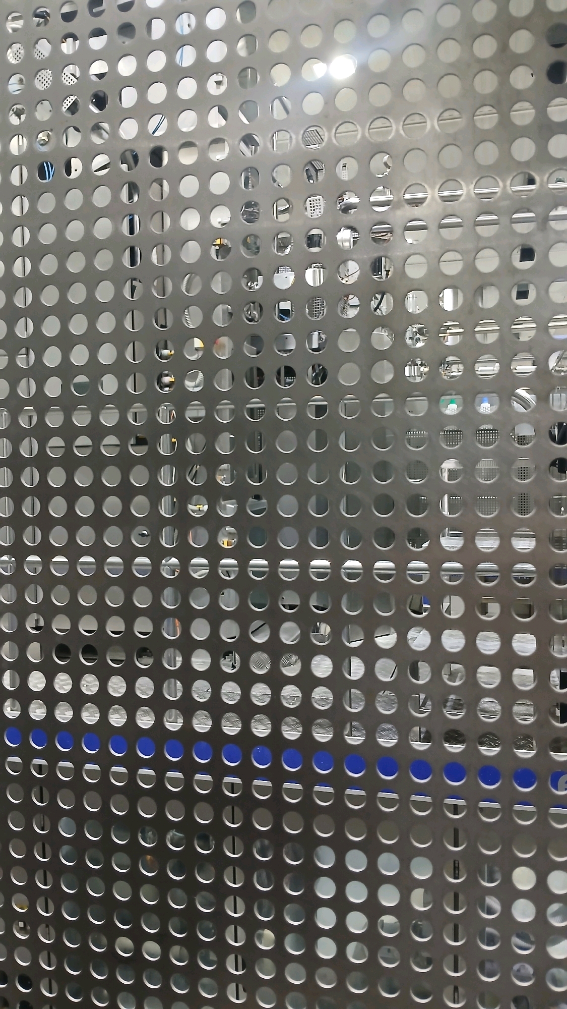 The image shows a close-up of a perforated metal sheet with a grid of circular holes. Some of the holes reveal machinery and equipment behind the sheet, suggesting an industrial or manufacturing setting. A few of the holes are filled with a solid blue color, creating a distinct visual pattern. The lighting in the image is bright, with reflections visible on the metal surface. The overall impression is of a functional and modern industrial space. There is no visible indication of the location being Gadobangkong, Indonesia, or any specific identifiable landmarks or people.