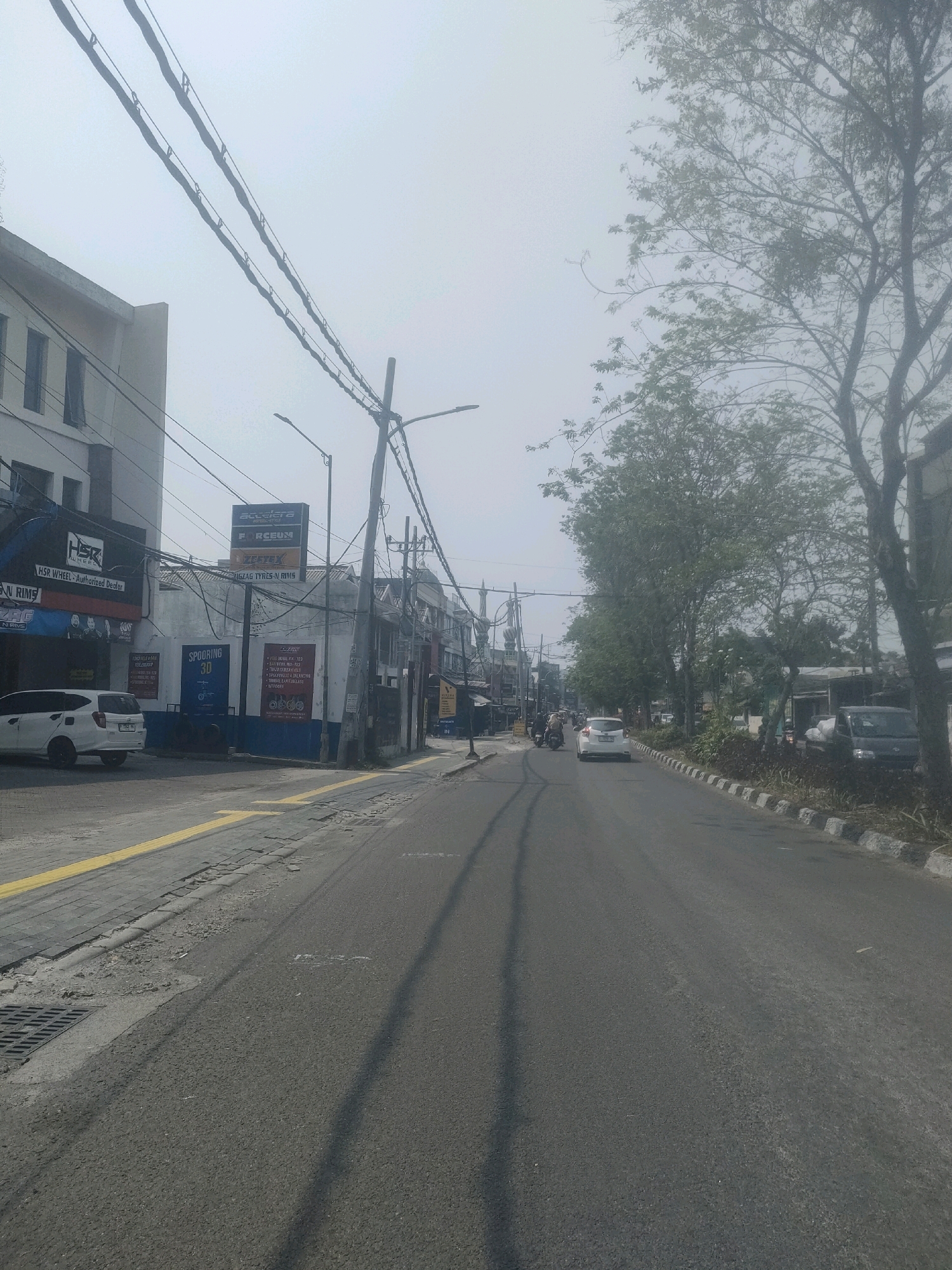 The image depicts a street scene in Surabaya, Indonesia, characterized by an overcast sky and a moderately built-up urban environment. On the left side of the road, a multi-story building with a light-colored facade is visible, featuring several windows. Adjacent to it, a commercial establishment with signage including "HSR WHEEL - AUTHORIZED DEALER" and "FORCEUN" is present. A white car is parked in front of this establishment. Further down the street, other businesses with various signs and awnings can be seen, along with a motorcycle and a car in motion. Power lines crisscross overhead, supported by poles. The road itself is asphalted, with a yellow line marking the edge of a sidewalk constructed with paving stones and concrete. On the right side of the road, trees with dense foliage line the street, interspersed with some vegetation and a darker-colored vehicle. The overall impression is a typical street in a busy Indonesian city, possibly an area with automotive-related businesses given the visible signage.