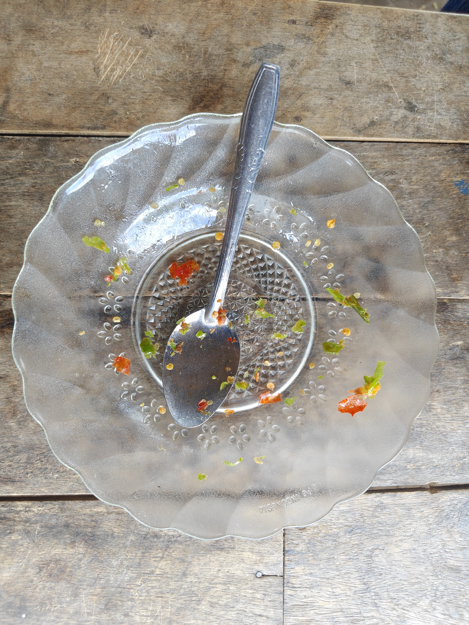 A dirty, clear glass plate with remnants of food, including pieces of chili and seeds, sits on a weathered wooden table. A silver spoon rests on the plate, also smeared with food residue. The table has visible grain and several scratches. The scene appears to be outdoors, possibly in a rural area, given the natural light and rustic setting.
