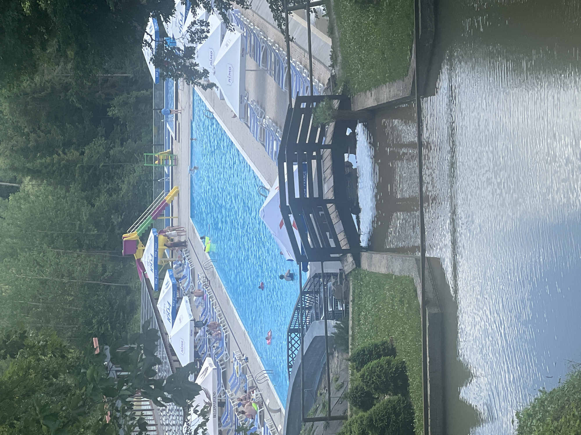 The image depicts a sunny day at a swimming pool complex, likely located in Khashchovania, Ukraine. The primary feature is a large, bright blue swimming pool filled with people. Several individuals are visible in the water, some swimming and others relaxing. In the foreground, a small wooden bridge spans a narrow waterway. Surrounding the pool, rows of empty lounge chairs are set up under large white umbrellas, suggesting a well-maintained recreational area. In the background, a vibrant waterslide complex with yellow and green slides adds to the playful atmosphere. Lush green trees and foliage frame the scene, indicating an outdoor setting. The overall impression is one of leisure and summer enjoyment.
