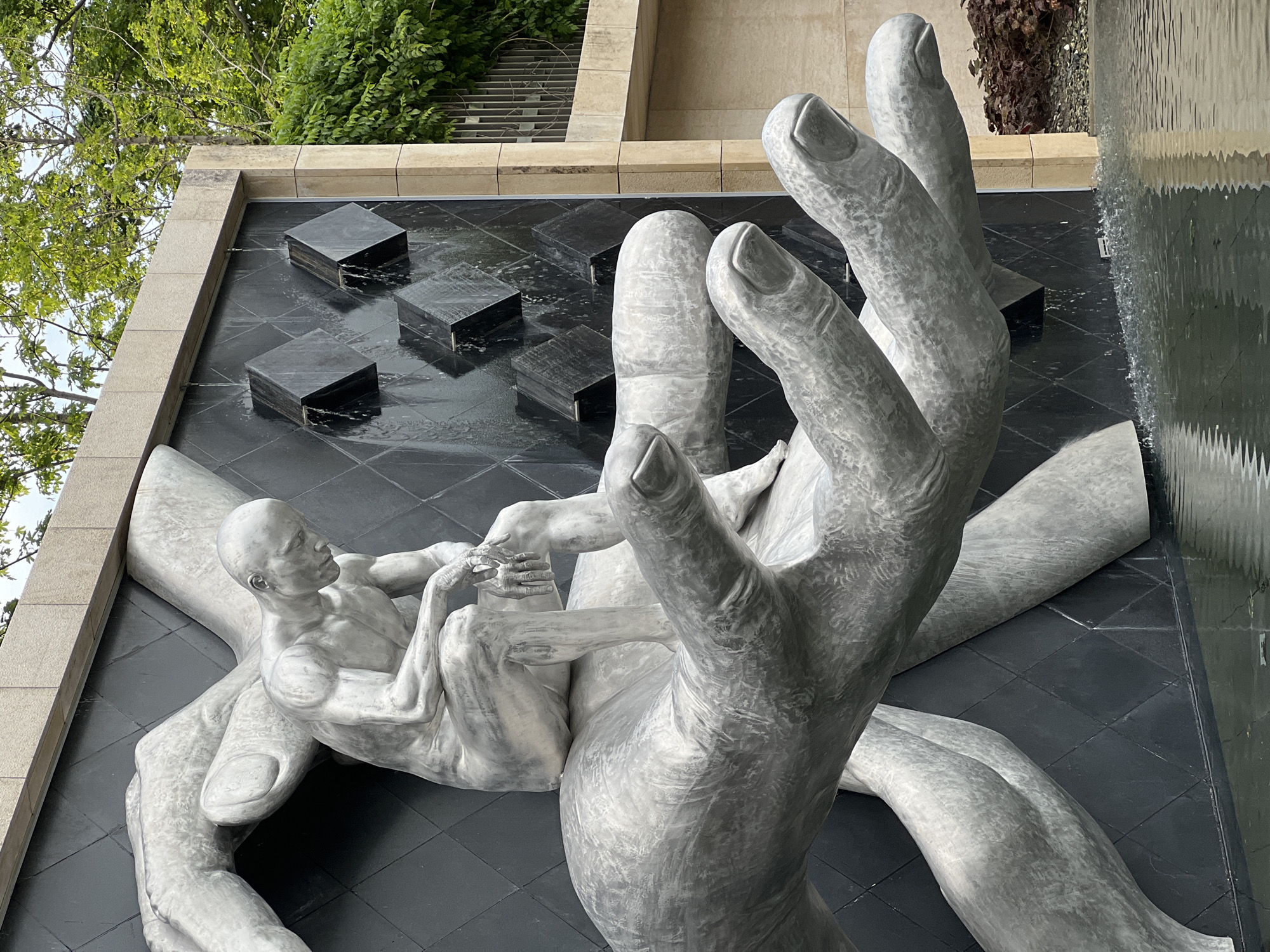 This image captures a striking outdoor sculptural water feature, likely part of a public garden or plaza in Krasnodar, Russia, given the provided context. The primary subjects are two oversized, textured gray hands, seemingly made of stone or concrete, which cradle a smaller, nude human figure of the same material.
The large hands dominate the foreground, with one prominent hand extending from the bottom right across the frame, its fingers subtly curled. The smaller, anatomically detailed male figure reclines within the larger hands, positioned on his back and looking upwards and slightly to the right. His left hand appears to gently touch one of the large fingers.
The sculptures are set within a dark, possibly black, tiled water basin. On the left side of the basin, several raised black square blocks feature small water jets that spray upwards, creating ripples on the dark surface. To the right, a continuous sheet of water cascades down a dark, reflective wall, creating a waterfall effect that contributes to the fountain's dynamic appeal.
The background reveals lush green foliage and tree branches at the top and upper left, suggesting an outdoor environment. Light-colored stone or concrete architectural elements frame the water feature, creating defined edges. The bright, even lighting indicates it is daytime. There is no visible text in the image.