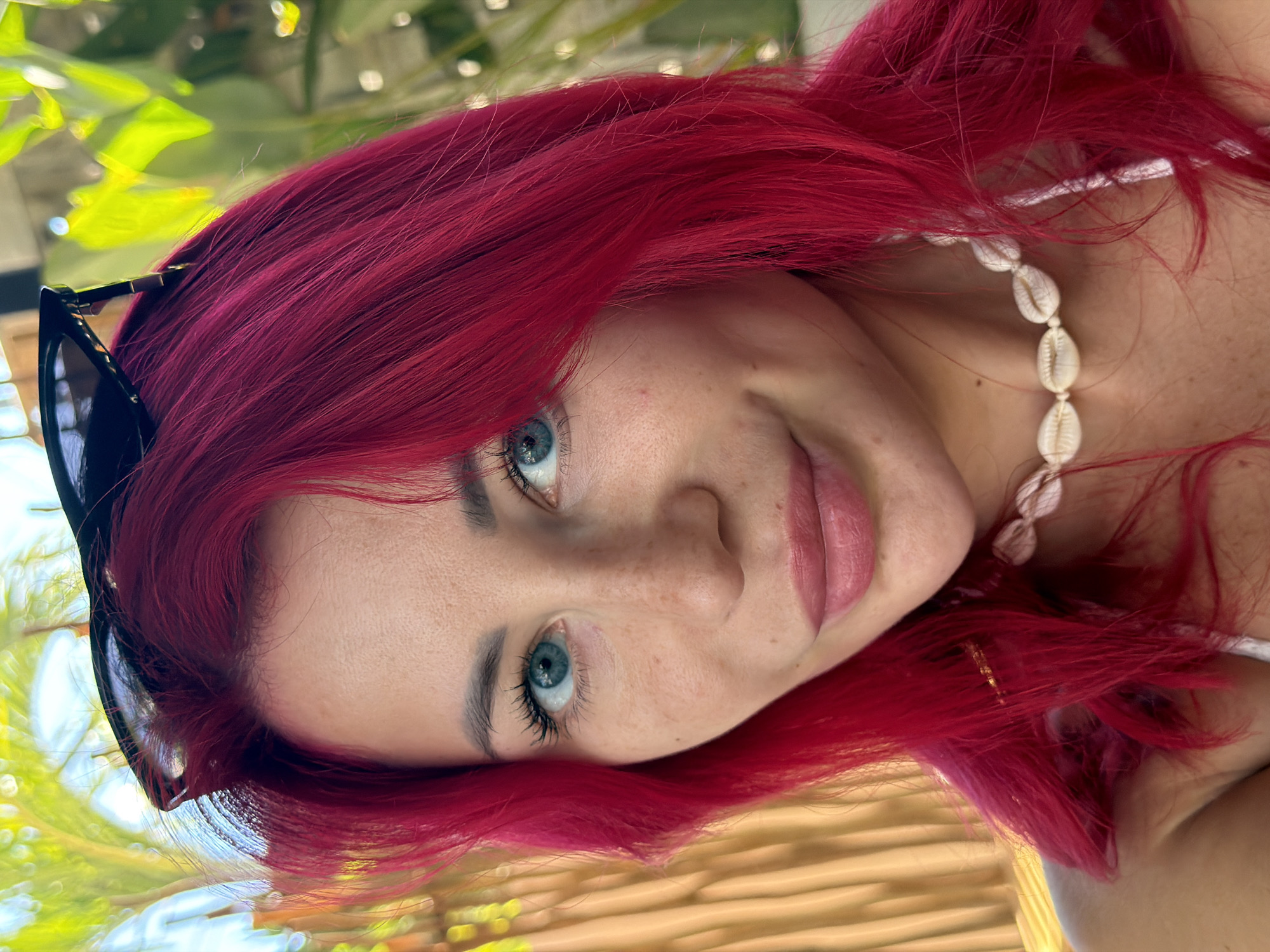 A woman with vibrant fuchsia hair and blue eyes smiles softly at the camera. She is wearing dark-rimmed sunglasses perched on her head, and a delicate seashell necklace. The background suggests an outdoor setting, with blurred greenery and warm-toned vertical blinds. The lighting is bright, indicating a sunny day. This scene appears to be captured in Kuta, Indonesia.