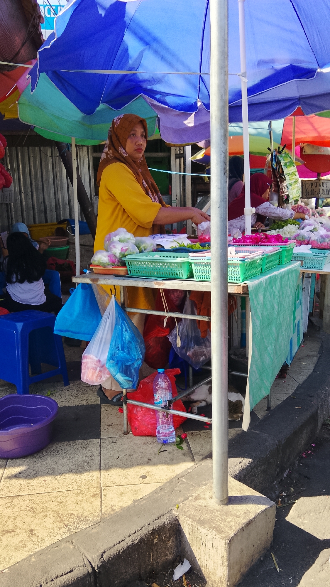 A street market stall is shown in Kemuteran, Indonesia. A woman wearing a yellow shirt and a brown hijab is standing behind a table laden with produce. She is partially obscured by a blue umbrella and a silver pole. The table holds several green baskets filled with produce. Bags of produce, plastic bags, and a red plastic bag are visible on the ground. A blue plastic chair is to the left of the stall. In the background, more market stalls and people are visible. The ground is paved with light-colored tiles.