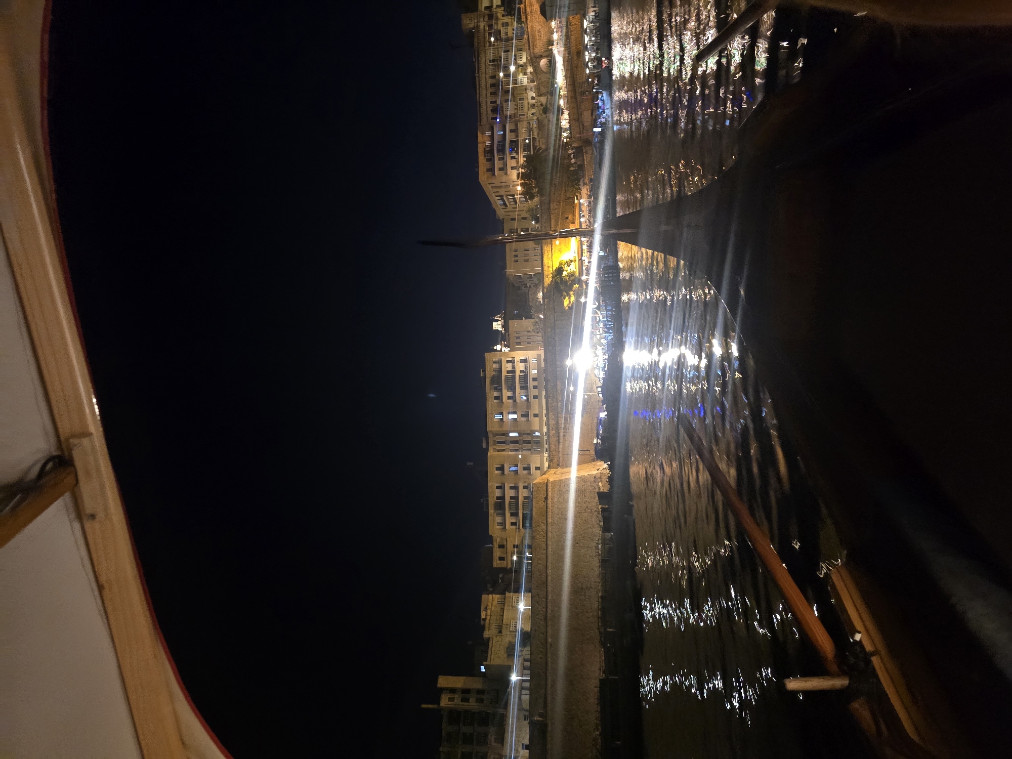 A nighttime view from a gondola in Valletta, Malta, shows the city's waterfront illuminated by streetlights and building lights reflecting on the water. The camera is positioned low, looking forward and slightly upwards, capturing the dark hull of the gondola in the foreground and two oars. In the mid-ground, a stone embankment lines the water, with buildings of various architectural styles, many with illuminated windows, situated behind it. The sky is dark, indicating it is nighttime. The scene suggests a leisurely boat ride along the harbor or a canal in Valletta, with the historic architecture of the city providing a backdrop.