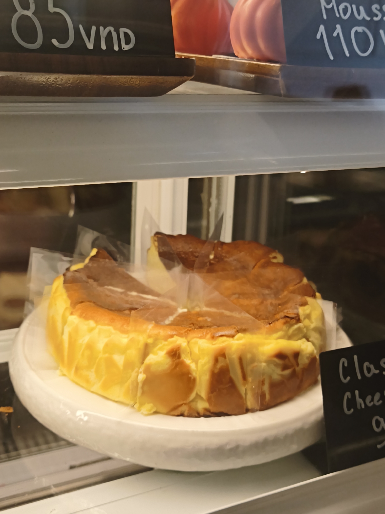 In the city of Nha Trang, Vietnam, within a bakery display case, a whole cheesecake is presented on a white cake stand. The cheesecake has a golden-brown, slightly cracked top and a yellow custard-like base. The cake is partially wrapped in clear plastic. To the right of the cheesecake, a small black sign with white chalk writing is visible, with the visible text reading "Clas Chees". In the background, a blurred reflection shows other items and the interior of the establishment. Above the cheesecake, two price tags are visible. One on the left reads "85 VNĐ" and the other, partially obscured, appears to read "110" with "Mousse" above it.