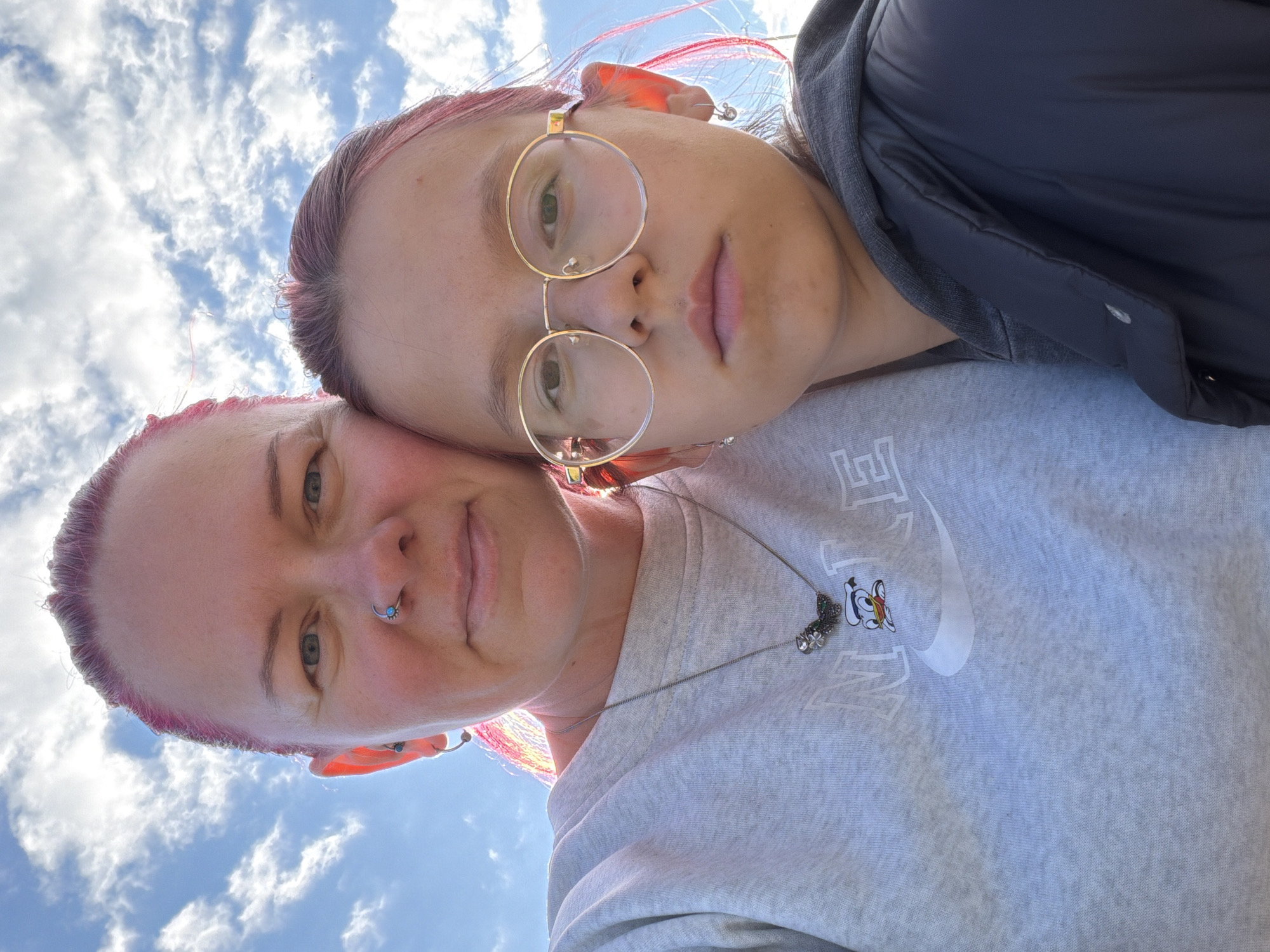 Two individuals, appearing to be an adult and a child, are captured in a selfie against a bright blue sky with scattered white clouds. The adult, on the left, has pink-tinted hair and is wearing a grey sweatshirt with a Nike logo and a Donald Duck graphic. A silver necklace with a butterfly charm adorns their neck. They also have a nose ring. The child, on the right, has round gold-rimmed glasses and also displays pink streaks in their hair. They are wearing a dark jacket over a grey hoodie. The lighting suggests it is daytime. The image does not contain enough information to determine a specific location within the city of Chaikovsky, Russia, or any other specific geographic details.
