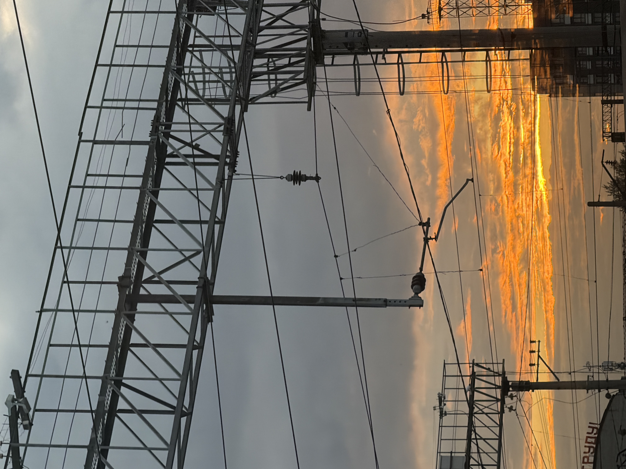 The image depicts an industrial scene, likely a railway yard, during a vibrant sunset in Perm, Russia. The sky is filled with dramatic orange and yellow clouds, casting a warm glow on the overcast sky. Overhead, intricate metal gantries support a network of electrical wires and insulators, typical of a railway infrastructure. These wires stretch across the frame, creating a complex web against the colorful sky. In the foreground and midground, sturdy concrete and metal poles support the overhead lines and associated equipment, including ladders and various electrical components. There are no people visible in the image. The overall impression is one of industrial utility set against a striking natural phenomenon.