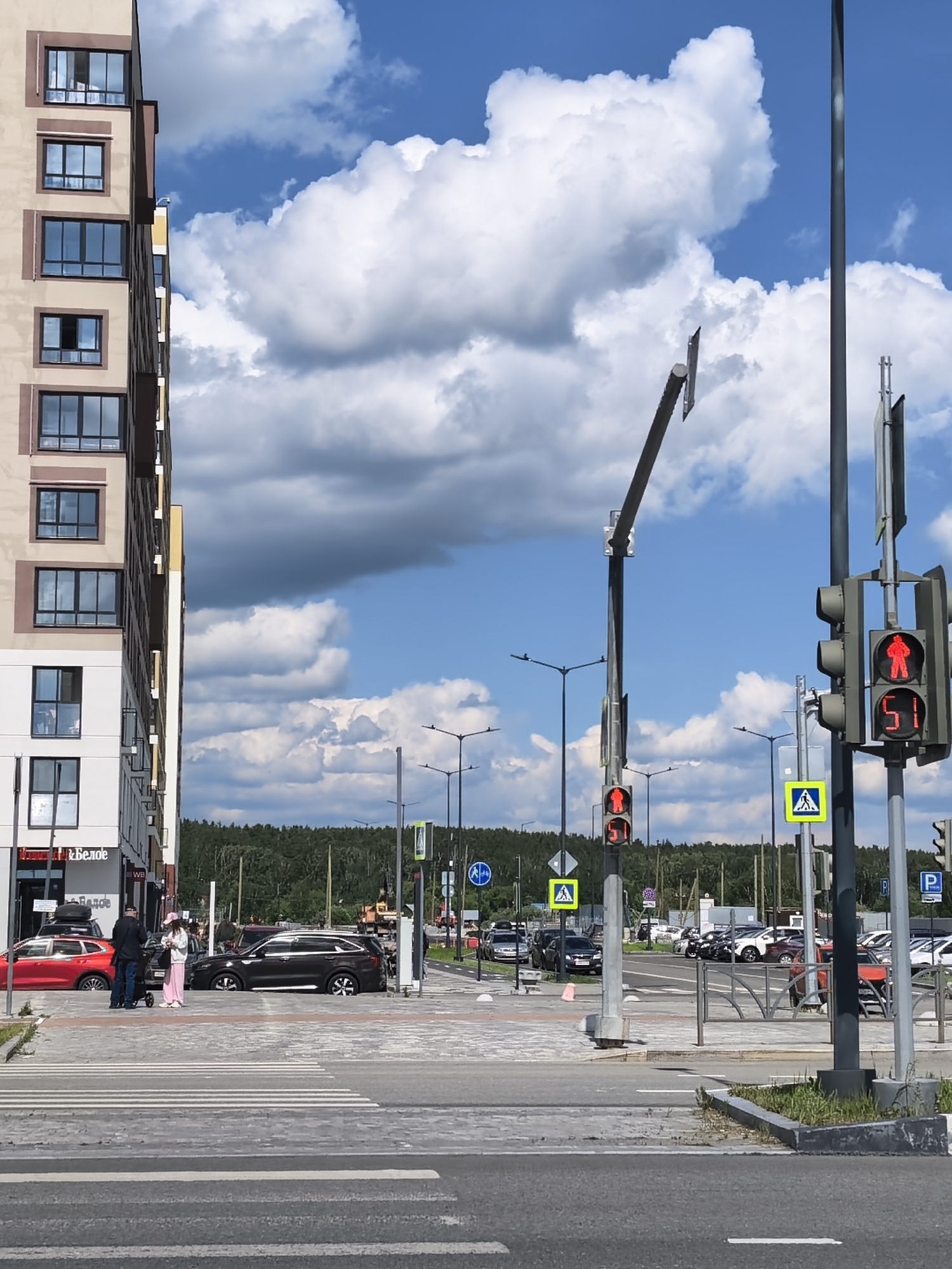 The image captures a city street scene in Unknown, Unknown on a sunny day with scattered clouds. A multi-story building stands to the left, featuring alternating tan and white facades with dark-framed windows. In the foreground, a zebra-striped crosswalk is visible. A family consisting of an adult male, an adult female, and a child in a stroller is standing on the sidewalk near the crosswalk. Several cars are parked along the street in the midground, and a line of trees can be seen in the distance. Traffic lights show red pedestrian signals, and various street signs are placed on poles near the crosswalk. Street lamps and other infrastructural elements are also visible.