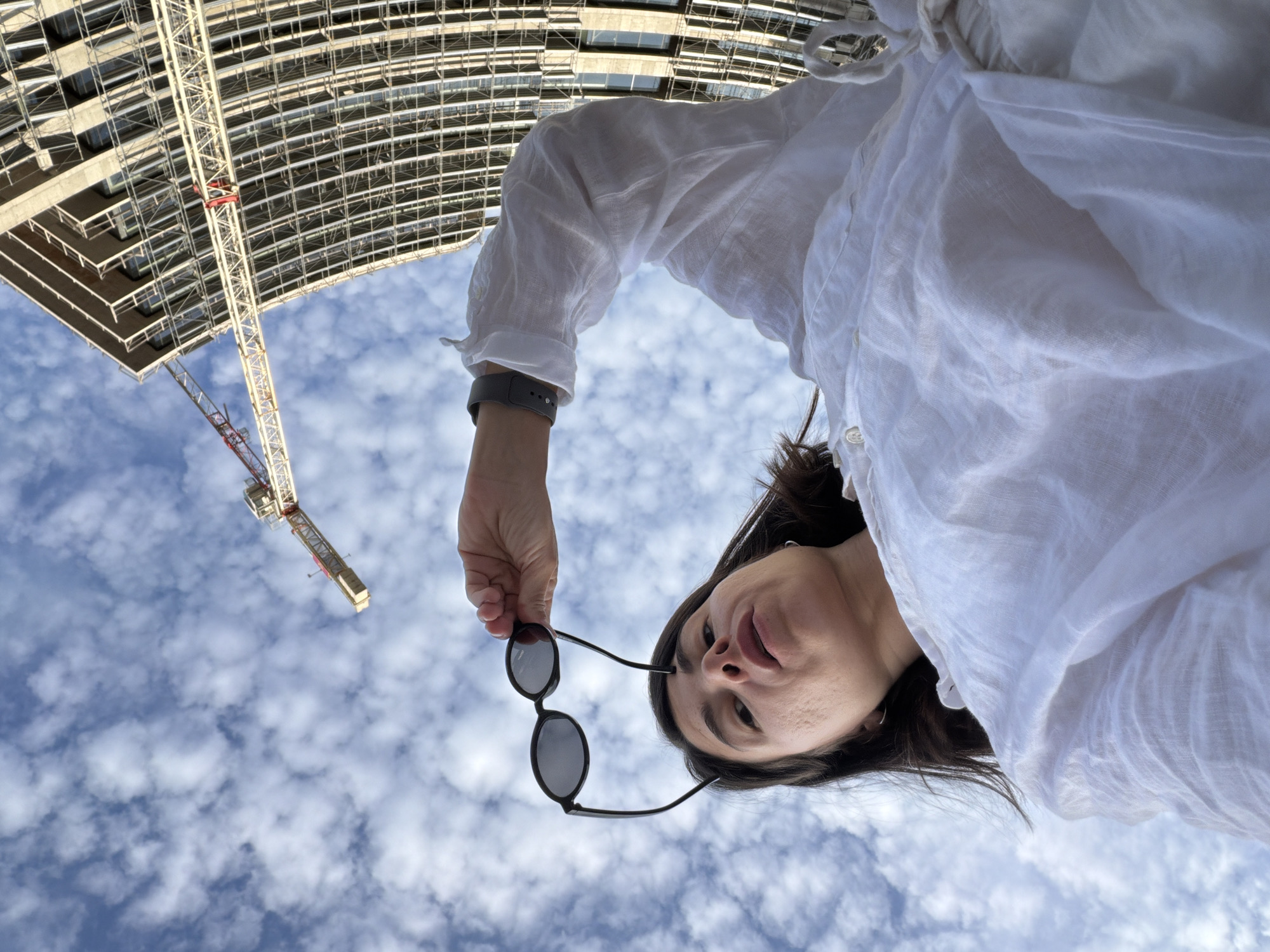 The image, captured in Budva, Montenegro, presents a low-angle perspective looking upwards. In the immediate foreground, an adult female is partially visible from the chest upwards, oriented towards the camera. She wears a white, loose-fitting linen shirt with visible buttons. Her dark hair frames her face, which is angled downwards, displaying a slightly open mouth. Her right hand is raised above her head, holding a pair of black-framed, dark-lensed oval sunglasses against the sky. A dark grey wristband is present on her left wrist. The background features a bright blue sky with numerous scattered white cumulus clouds. To the right, a multi-story building under construction is visible, characterized by an exposed concrete framework and a grid-like pattern of scaffolding or structural elements. A red and white construction crane extends from the left side of the frame, its boom partially visible against the sky, positioned in proximity to the building.