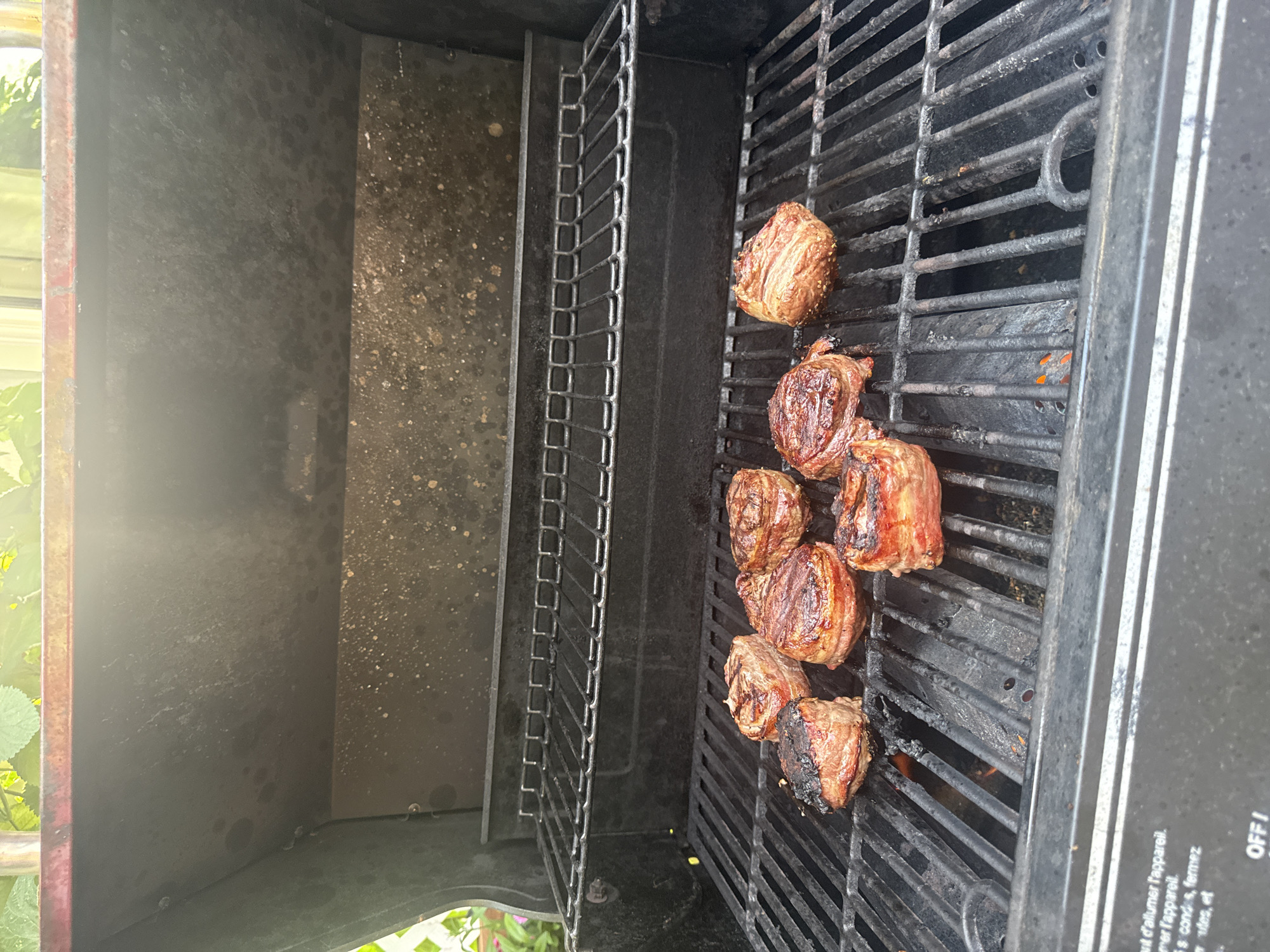 In Calgary, Canada, a close-up, eye-level shot shows several bacon-wrapped steaks cooking on the grates of a black gas grill. The steaks, which appear to be thick-cut medallions, are arranged on the lower grate of the grill. Some steaks show distinct grill marks and a medium-rare to medium doneness, while one steak on the far left is slightly more charred. The grill itself appears well-used, with visible grease stains and some debris on the interior surfaces and grates. A second, higher grate is visible directly above the steaks. The background shows blurred greenery, suggesting an outdoor setting. The bottom of the image reveals the control panel of the grill, with some faded French text that appears to be instructions for lighting the appliance.