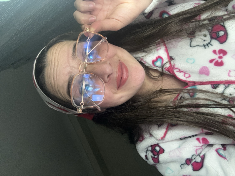 A woman with long brown hair and a headband holds up gold-framed eyeglasses. She is wearing a white pajama top with a Hello Kitty pattern. She looks directly at the camera with a slight smile. The background is dimly lit, suggesting an indoor setting. The image is rotated, giving a tilted perspective.