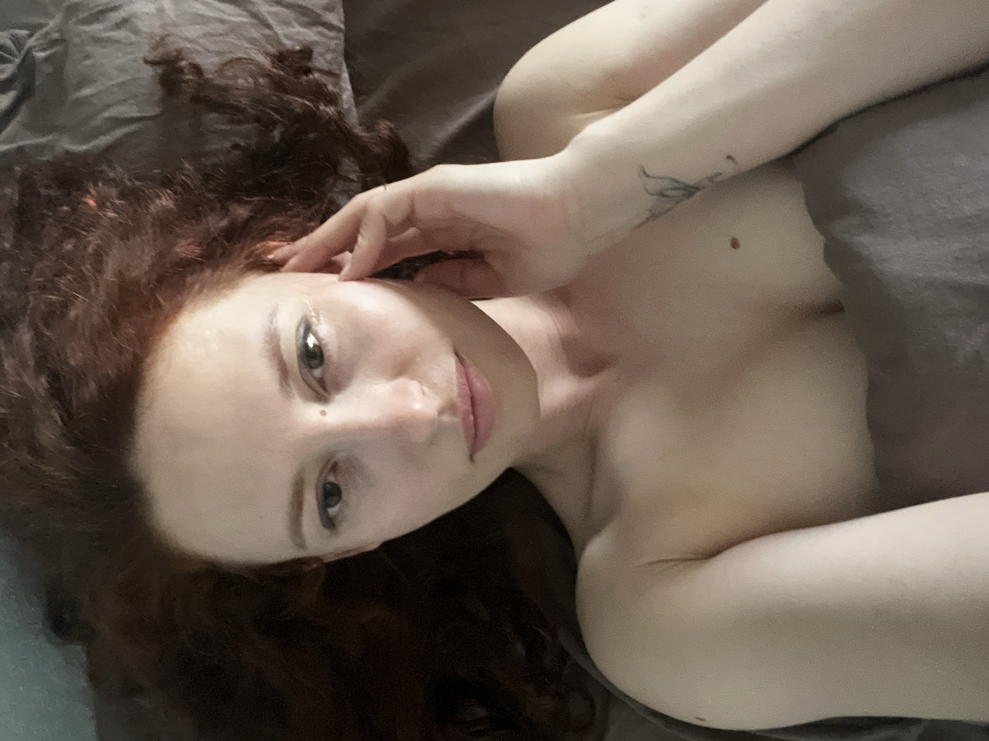 A fair-skinned person with red, curly hair is lying in bed. The person's head is resting on a grey pillow or blanket. Their right arm is raised with the hand near their face. The person is looking at the camera. A tattoo is visible on the inner left forearm. The location cannot be determined.