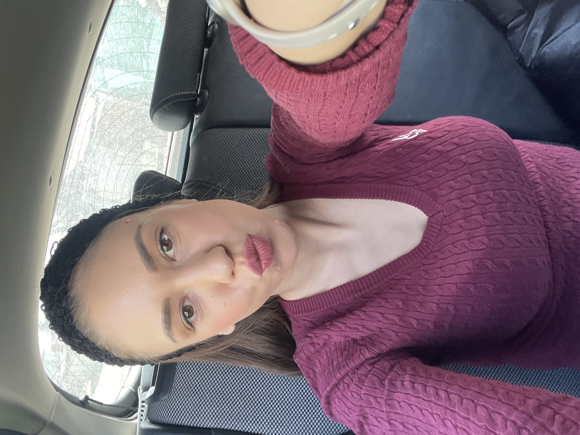 A selfie was taken inside a car, in Dobrogled, Bulgaria. A female subject with brown hair is wearing a black headband and a maroon sweater with a low-cut neckline. The interior of the car includes black seats, a headrest, and a view through the rear window obscured by condensation or frost.