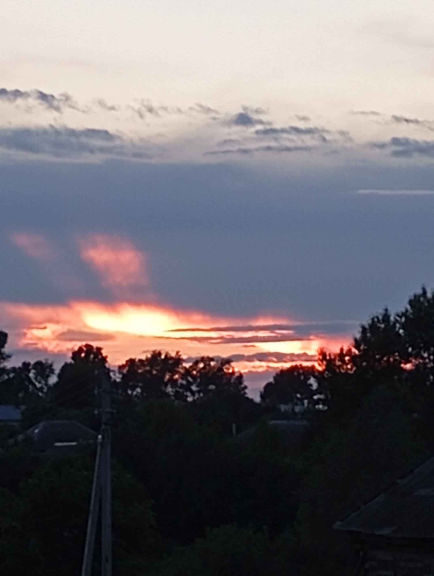 The image captures a twilight scene over the city of Foky, Russia. The sky is dominated by a dramatic sunset, with hues of orange and pink bleeding through a band of dark clouds. Below the horizon, a silhouette of trees and the roofs of buildings are visible against the darkening sky. A utility pole stands prominently in the foreground on the left side of the frame. The overall impression is one of a serene, end-of-day landscape.