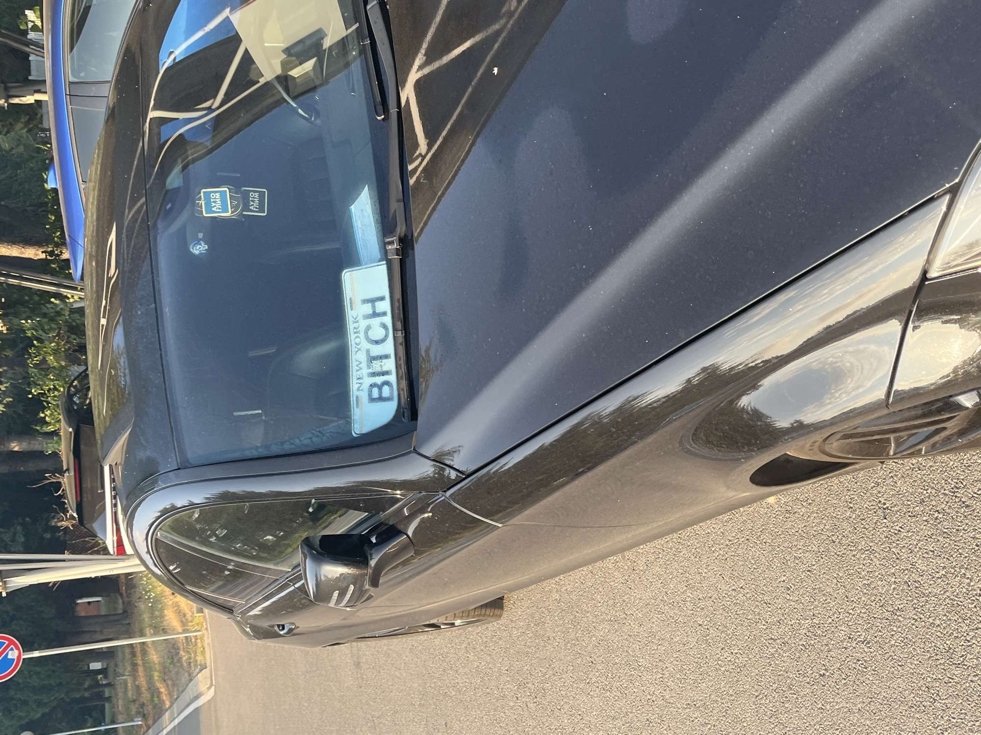 A black Mercedes-Benz CLS is parked on an asphalt road in Nesebar, Bulgaria. The car has a custom license plate on the windshield that reads "NEW YORK BITCH". There is an air freshener hanging from the rearview mirror. In the background, other vehicles and trees are visible. The sun is shining, casting reflections on the car's surface.