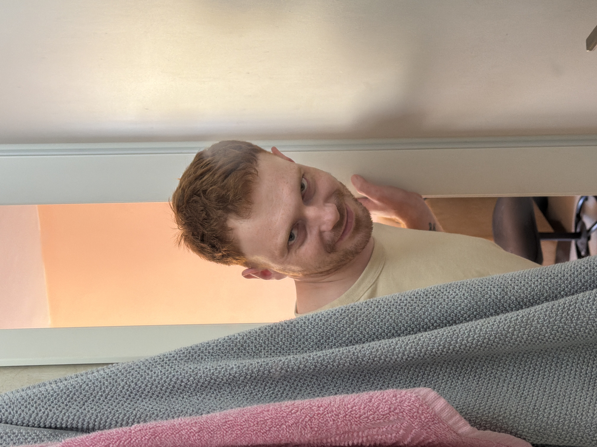 A man with red hair and a beard is peeking from behind a door frame. He is wearing a light yellow t-shirt and has his left hand on the door frame. Two towels, one pink and one gray, hang to the left of the frame. The background shows a room with an orange-toned wall and a chair with a black seat. The image was taken in Tel Aviv, Israel.