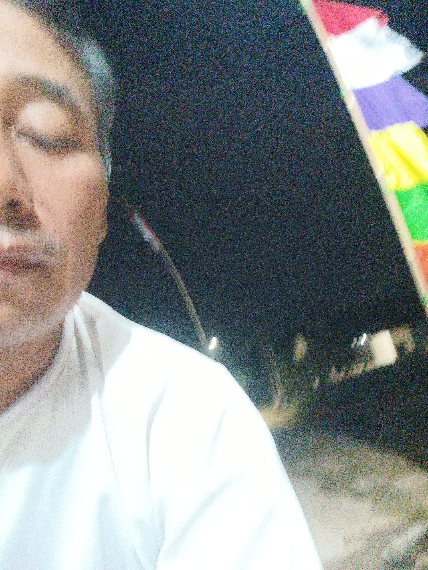 A close-up, low-light, slightly grainy photograph taken from the chest up features a person's face on the left side of the frame. The individual, appearing to be male, has his eyes closed and is wearing a white shirt that occupies the majority of the lower right portion of the image. To the right of the person's head, a series of colorful, triangular flags are visible, strung together and angled upwards. The background is dark, suggesting it is nighttime, with some blurry lights and foliage discernible in the distance. The image was captured on Jalan Tegal Gondo, Indonesia.