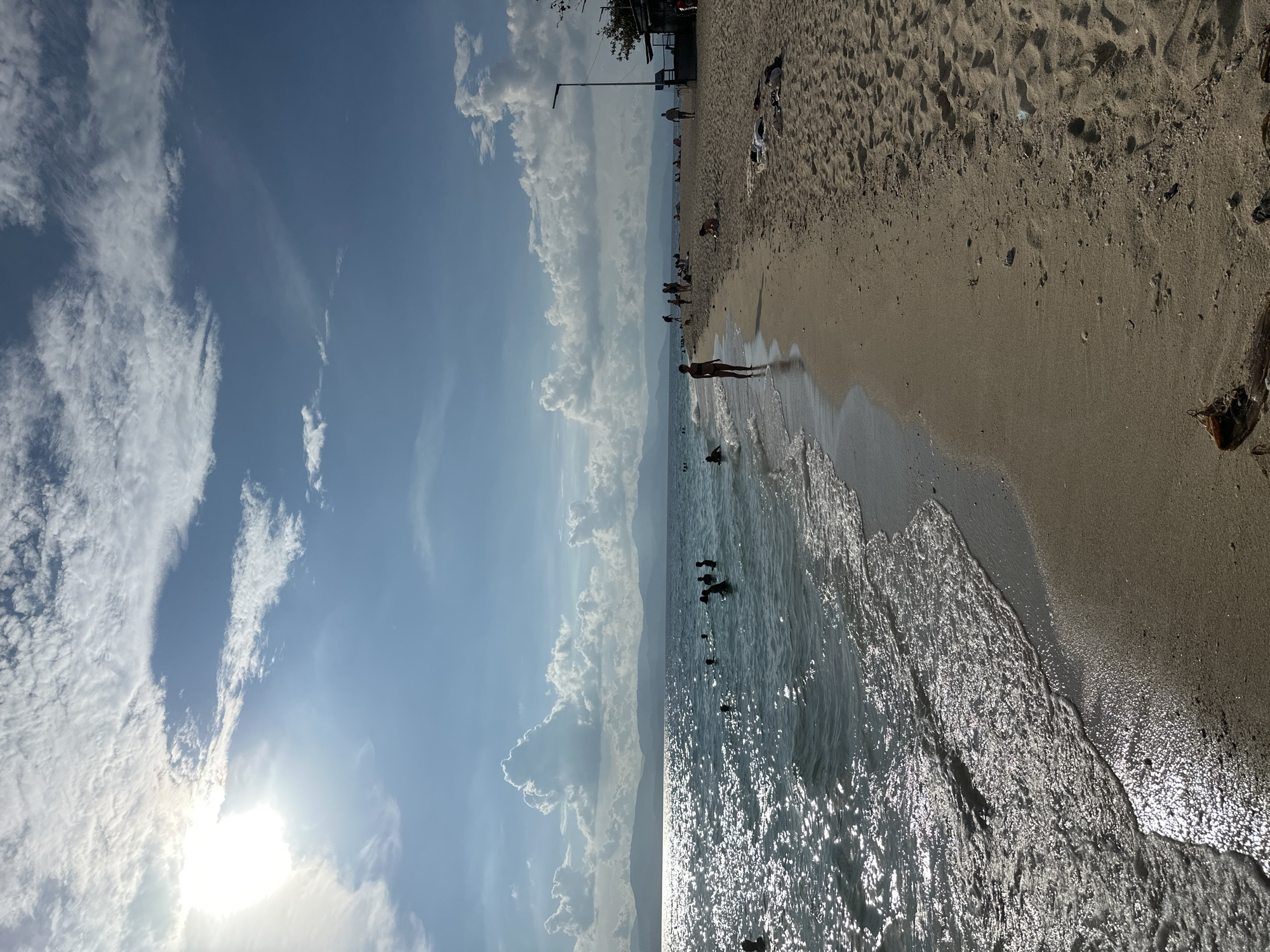The image depicts a sunny day at a beach in Saavedra, Philippines, with the sun high in the sky and bright sunlight reflecting off the water. The sea is calm with small waves lapping at the sandy shore. Several people are visible in the water, swimming or wading, and on the beach, some are walking, sitting, or lying down. In the background, mountains form a distant silhouette against the horizon. The sky is partly cloudy with white cumulus clouds. A few structures, including a lamppost and what appears to be a small building or kiosk, are present on the right side of the beach. Debris, such as a piece of driftwood, is scattered on the sand.