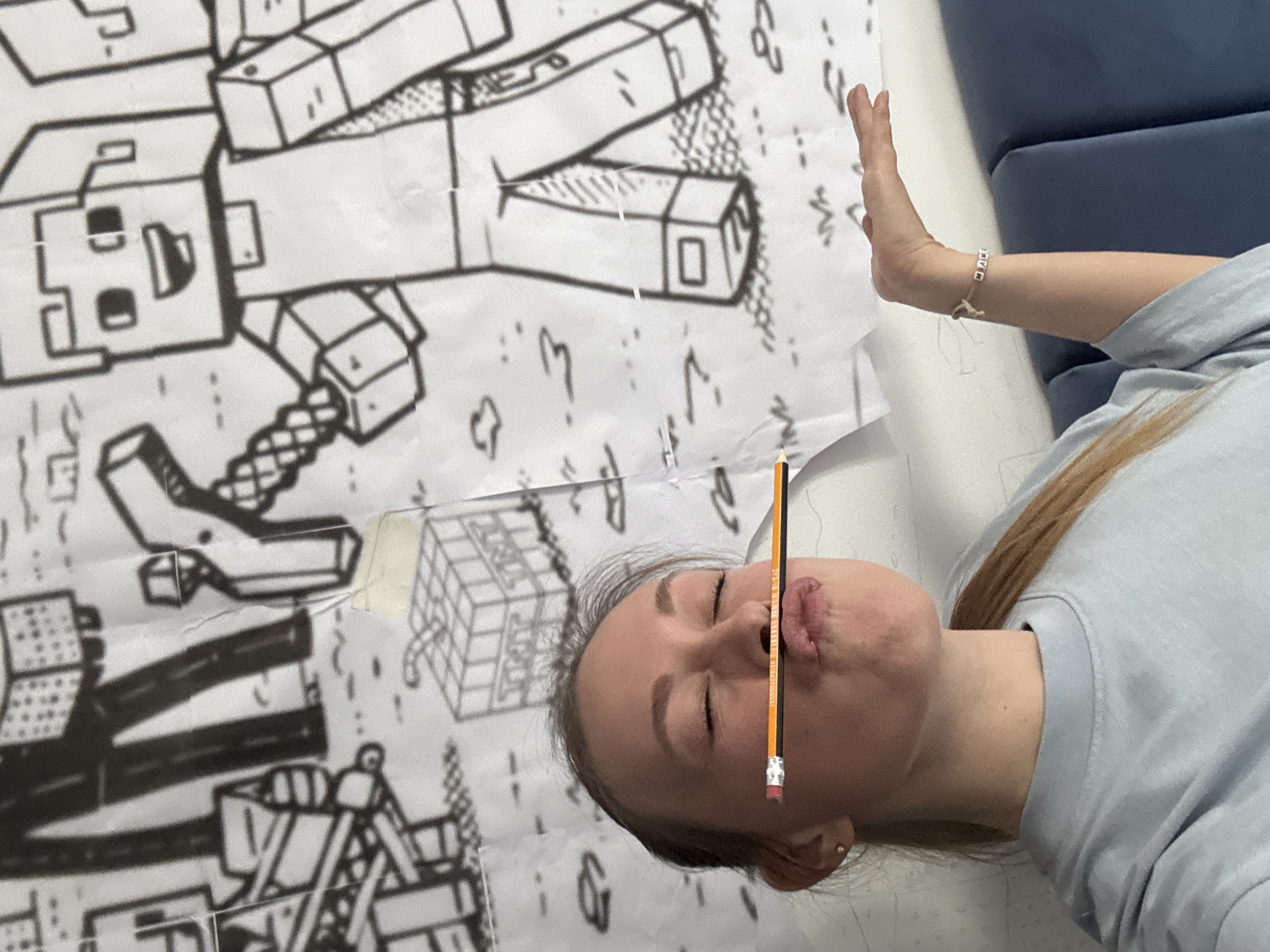 A woman is lying on a blue couch and holding a pencil in her mouth. She is wearing a light blue shirt and a bracelet on her right wrist. Behind her is a large sheet of paper with black and white line drawings that appear to be architectural or geometric in nature. The location is unknown and is not determinable from this image.