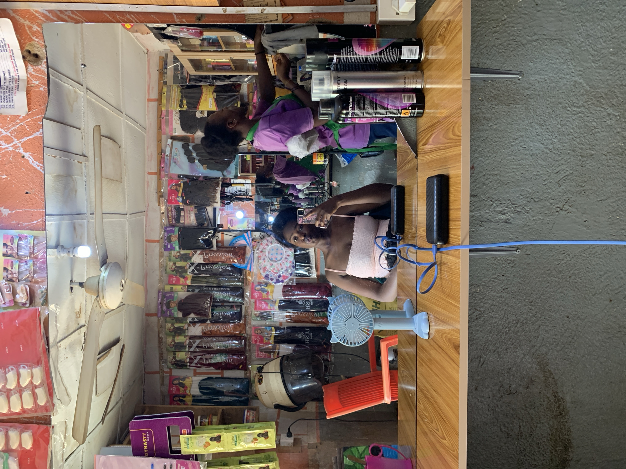 The image shows the interior of a small retail shop in Rafin Par, Nigeria. The shop sells hair products, extensions, and beauty accessories. A young woman with short dark hair is taking a selfie in the shop mirror. She is wearing a light pink tank top. Behind her, on shelves, are multiple packages of hair extensions. A small blue electric fan and a hair dryer are on a counter next to her. Above her in the mirror, another woman in a purple shirt and green apron is reaching for hair products. Several cans of hair spray are lined up on the counter. A ceiling fan is visible in the left side of the frame. Printed price lists are on the wall.