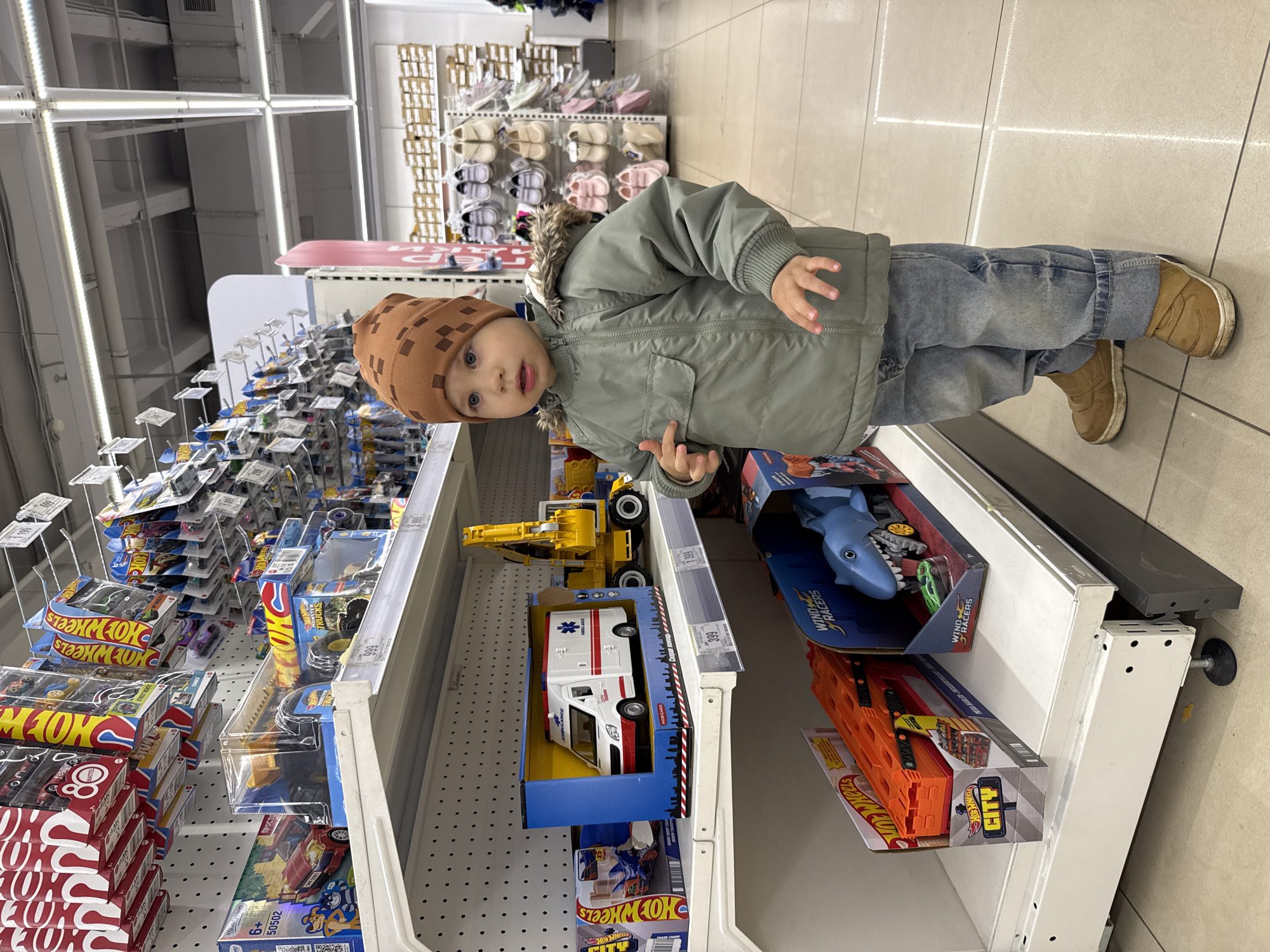 The image shows a young child standing in front of a shelf in a store, examining toys. The child is wearing a brown patterned hat, a green jacket with a fur-trimmed hood, blue jeans, and brown shoes. The shelf is stocked with various toys, including Hot Wheels cars and trucks, a toy ambulance, and a shark-themed toy. The location is likely a retail establishment in Korobovo, Russia, based on the presence of items and labels written in languages commonly found in that region. The child is looking at the camera with a slightly inquisitive expression. The environment appears to be a well-lit store aisle.