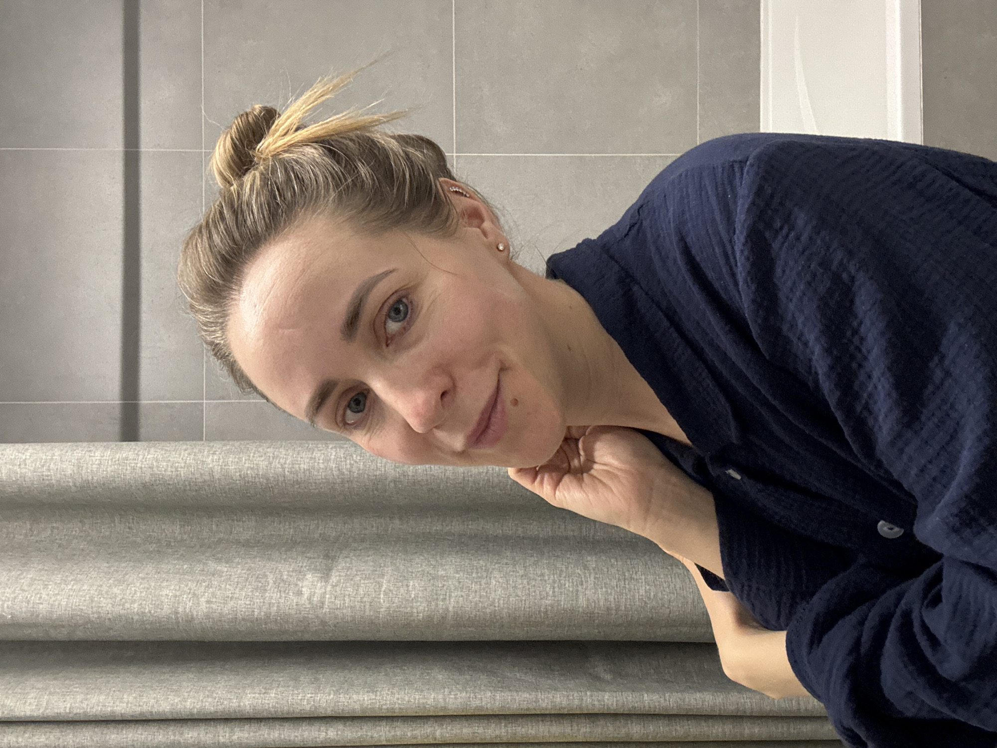 An indoor portrait displays a young adult woman looking directly at the camera with a gentle, soft smile. Her head is tilted, and her chin rests on her right hand, which is lightly curled. She has light skin, blue eyes, and blonde to light brown hair pulled back into a high, messy bun, with some strands appearing lighter and more golden. She wears small stud earrings in her earlobes and a delicate helix piercing featuring a line of tiny, clear stones. A faint beauty mark or mole is visible on her right cheek/jaw area.
She is dressed in a dark blue, possibly navy, top or robe made of a textured, crinkled fabric, possibly muslin or gauze, with visible buttons on the front.
The background consists of light grey tiled walls with horizontal and vertical grout lines in the upper section, suggesting a modern bathroom or utility space. In the lower portion of the image, light grey fabric blinds or Roman shades are visible, folded horizontally in several layers. A narrow white vertical structure, possibly part of a door frame or cabinet, is partially visible in the upper right.
The lighting is soft and even, indicating either controlled artificial light or diffuse daylight. The woman's expression is calm and inviting. There is no visible text in the image. The setting is in Chaikovsky, Russia.