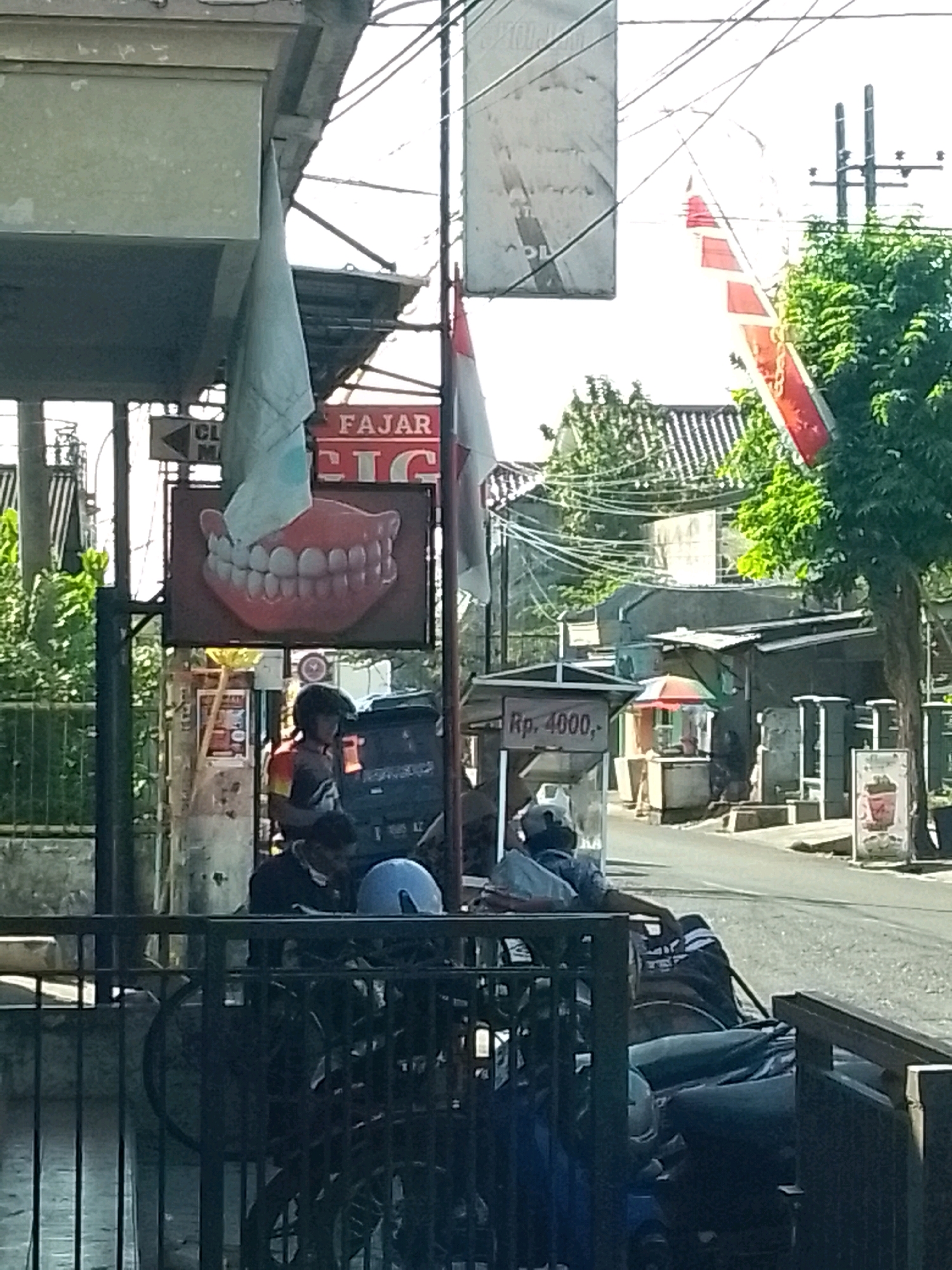 The image depicts a street scene in Gebang, Indonesia, under bright daylight conditions. In the foreground, a black metal fence borders a sidewalk. Behind the fence, several motorcycles are parked. One motorcycle in the center has a person in a helmet sitting on it, wearing a multi-colored shirt. Another person in a blue helmet is partially visible in the lower right portion of the frame.
Further back, a prominent sign advertising "FAJAR CIG" with a large graphic of human teeth is visible. To the left of this sign, a smaller sign with a triangular arrow and text starting with "CL" is present. A light blue flag hangs from a pole near the "FAJAR CIG" sign.
To the right of the "FAJAR CIG" sign, a street vendor's cart is set up, displaying the price "Rp. 4000,-" on its awning. A small red and white umbrella is open above the cart. In the background, a street stretches into the distance, lined with buildings and trees. Electrical wires are strung overhead. The overall impression is a bustling commercial area with street vendors and parked vehicles.