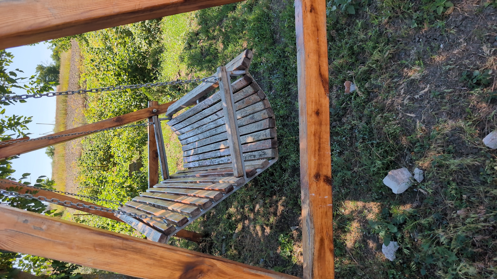 A wooden swing set with a slatted wooden seat is positioned in a grassy area. The swing seat is suspended by metal chains from the wooden A-frame structure. In the background, there is a field with green vegetation and some distant trees under a clear sky. The ground around the swing set is covered in grass and small stones. The image appears to be taken outdoors during daylight.