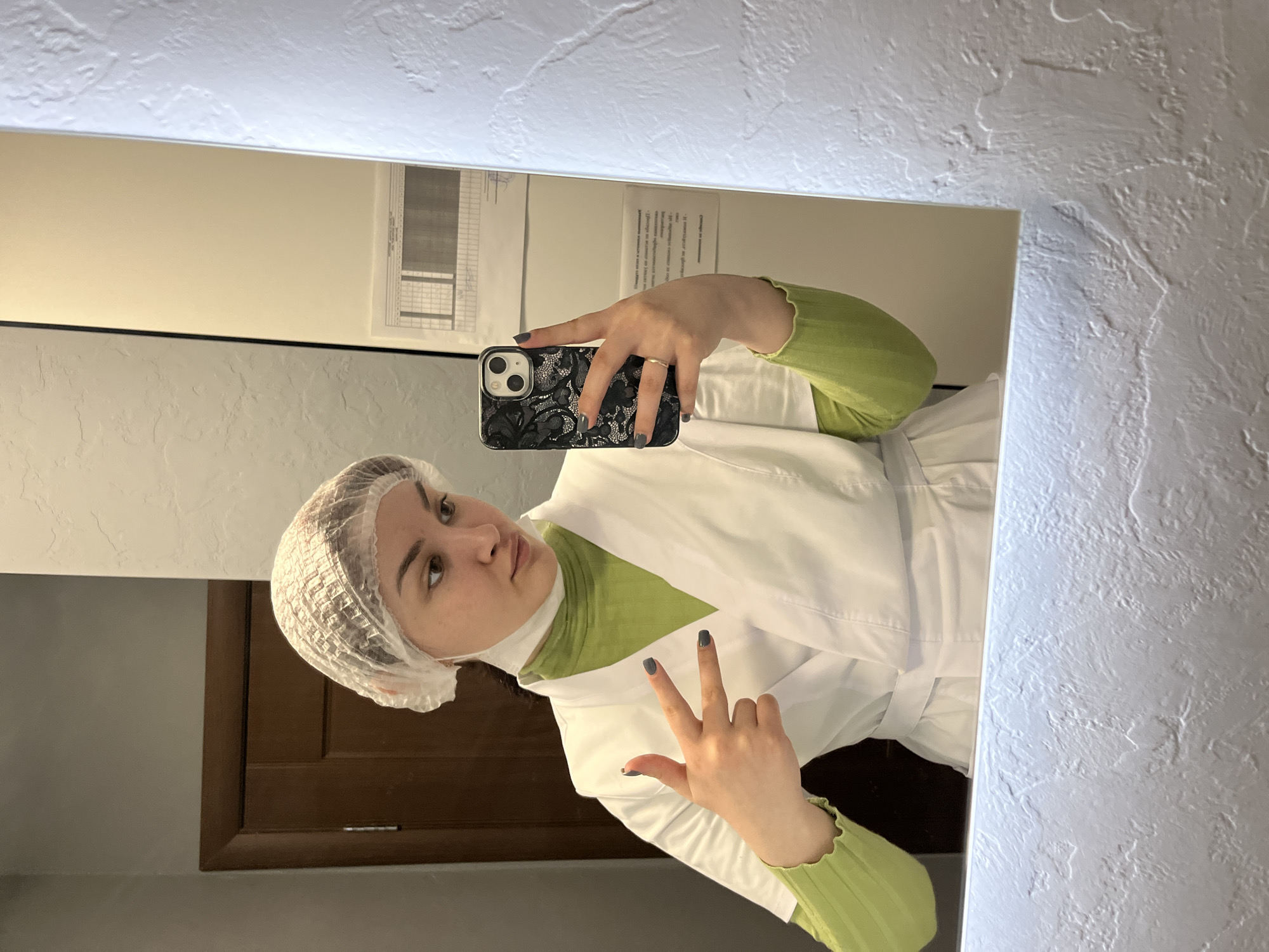 A woman is taking a selfie in a mirror. She is wearing a white lab coat, a green shirt, and a white hair net. She is holding a black phone with a patterned case. She is making a peace sign with her hand. The background shows a plain white wall with a brown cabinet or door visible in the lower left corner. Above her, there is a document or chart hanging on the wall. The environment appears to be a clinic or laboratory, though the specific location in Neftekamsk, Russia, is not discernible from the image alone.